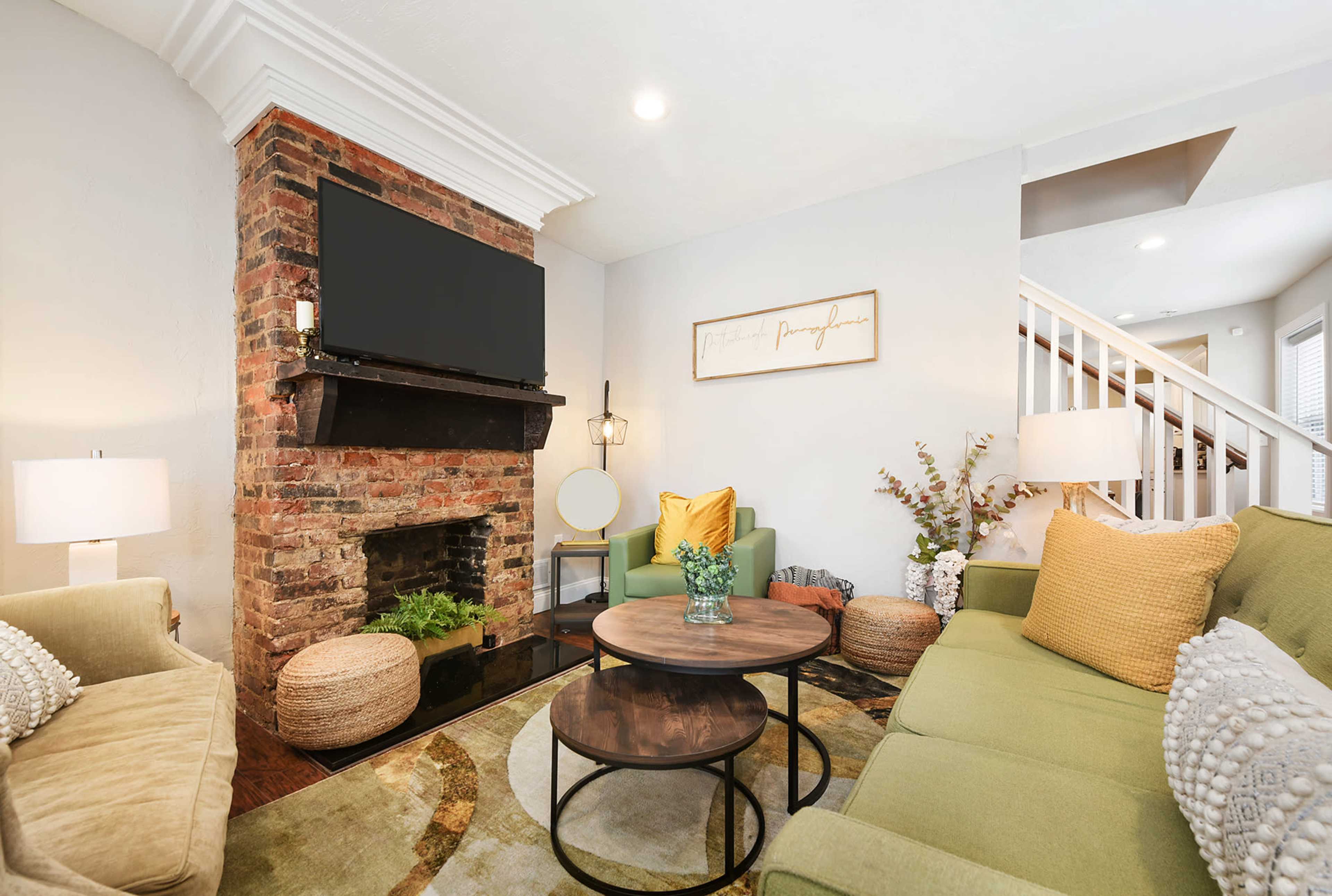 The living room features a brick fireplace with a mounted television, surrounded by green and yellow seating, a circular coffee table, and decorative plants.