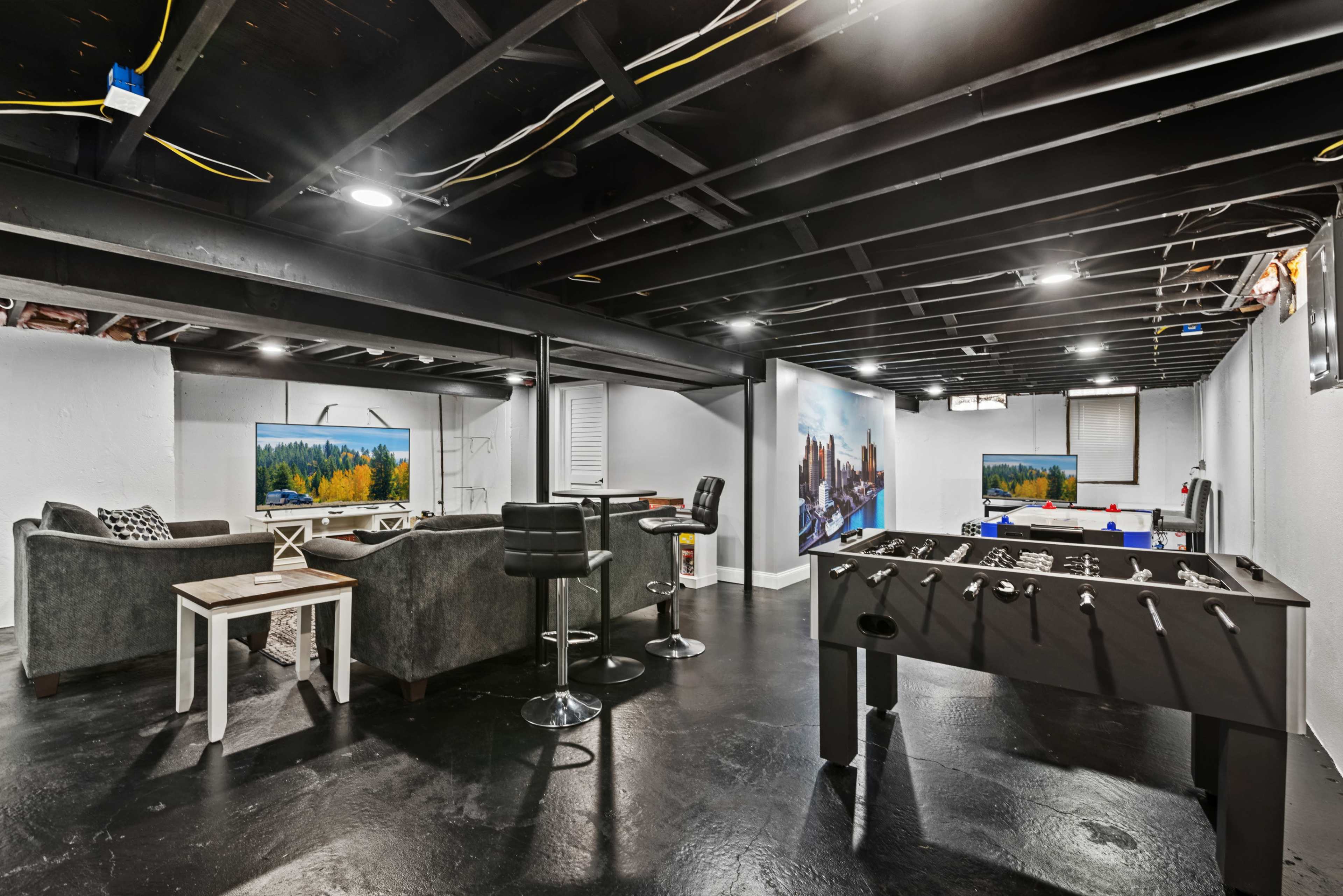 A modern basement recreation room with gray sofas, a foosball table, and large wall-mounted televisions displaying scenic images.