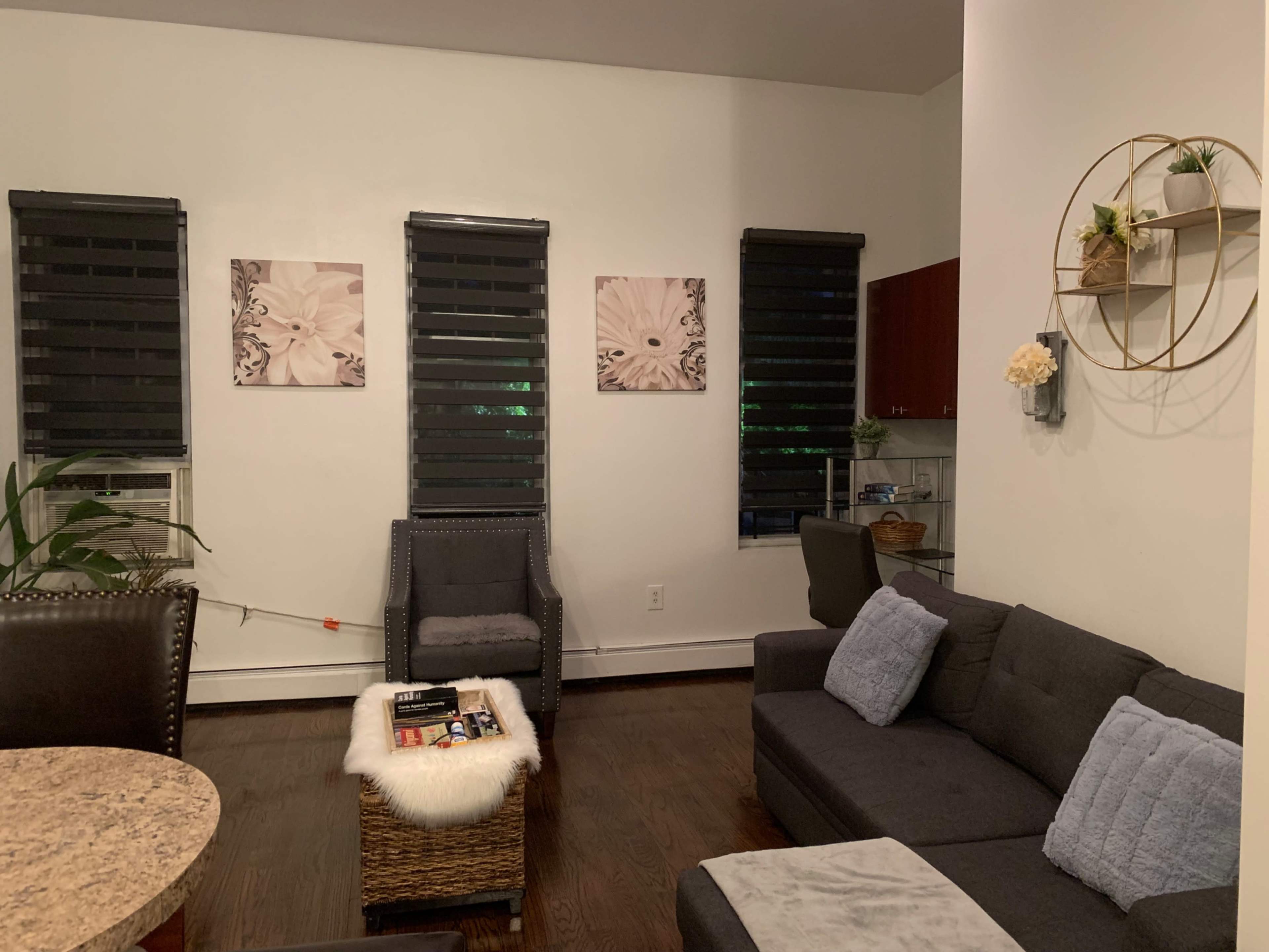 A living room with dark wood flooring, a gray sectional sofa, two framed floral pictures on the wall, and a small shelf with decorative items.