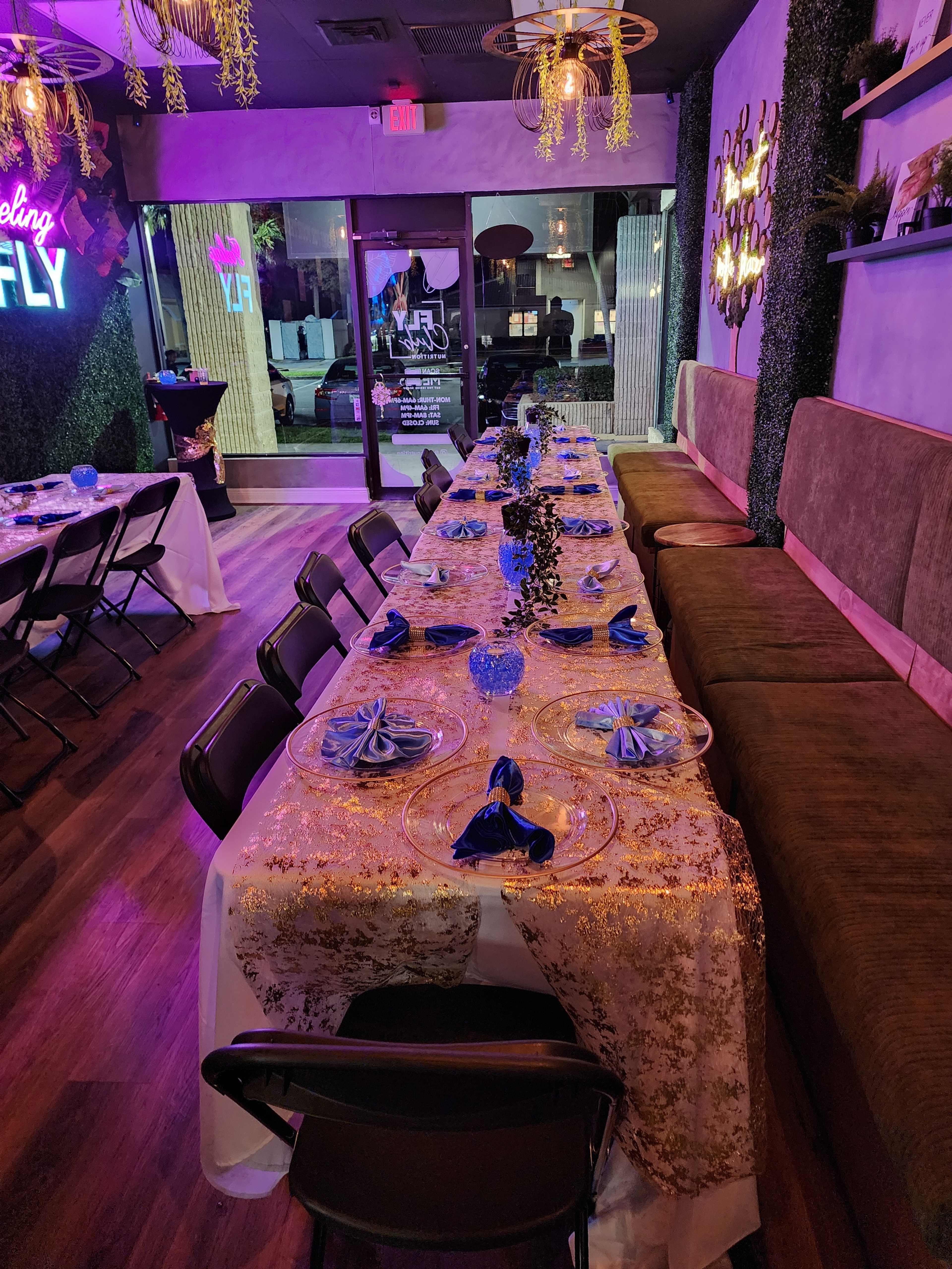 The image shows a decorated dining table set up in a restaurant with blue napkins, plates, and a golden table runner, alongside a cozy seating area.