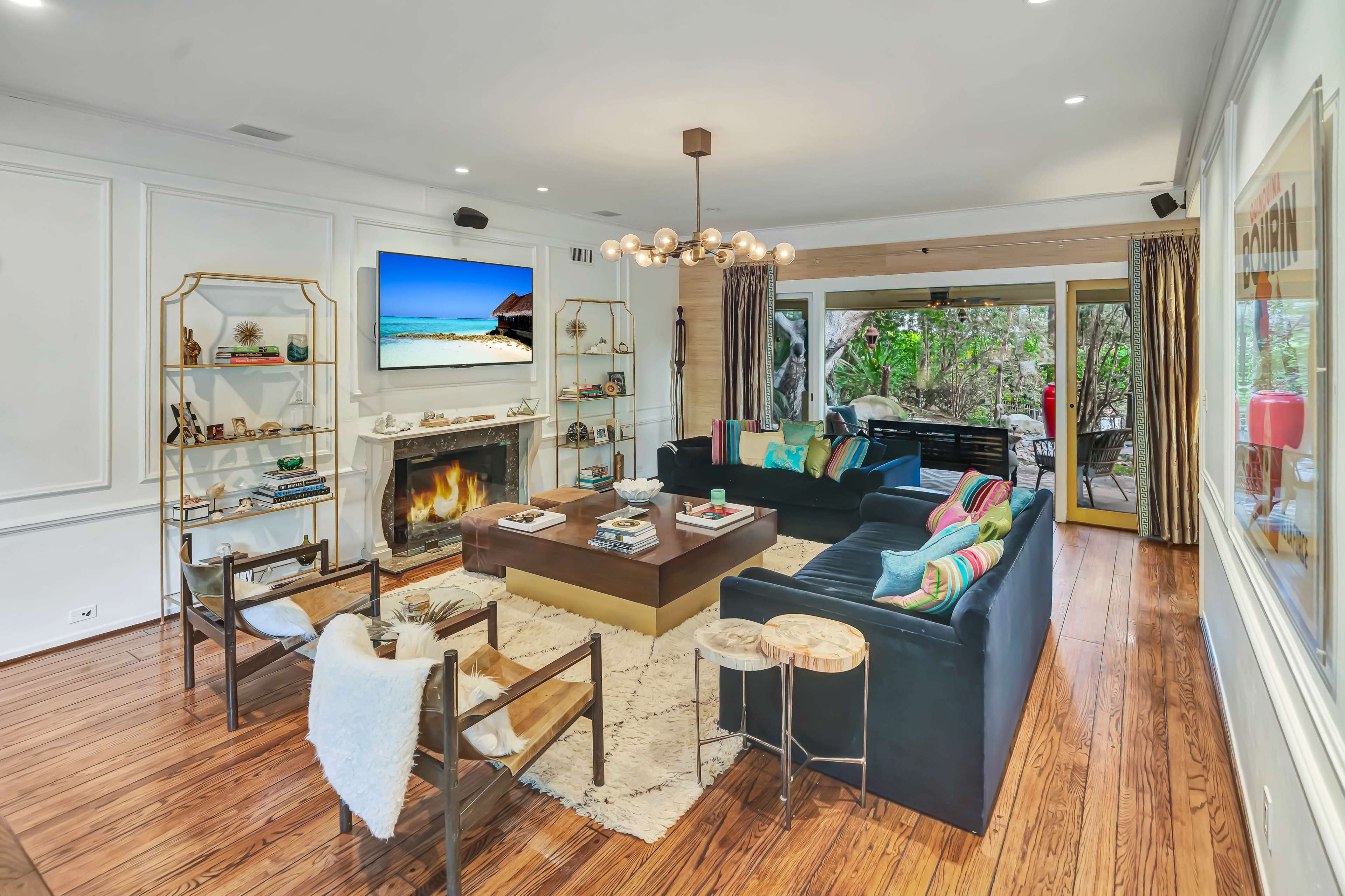 The image shows a modern living room with a central coffee table, comfortable seating arrangements, a large television displaying a beach scene, and access to a patio through glass doors.