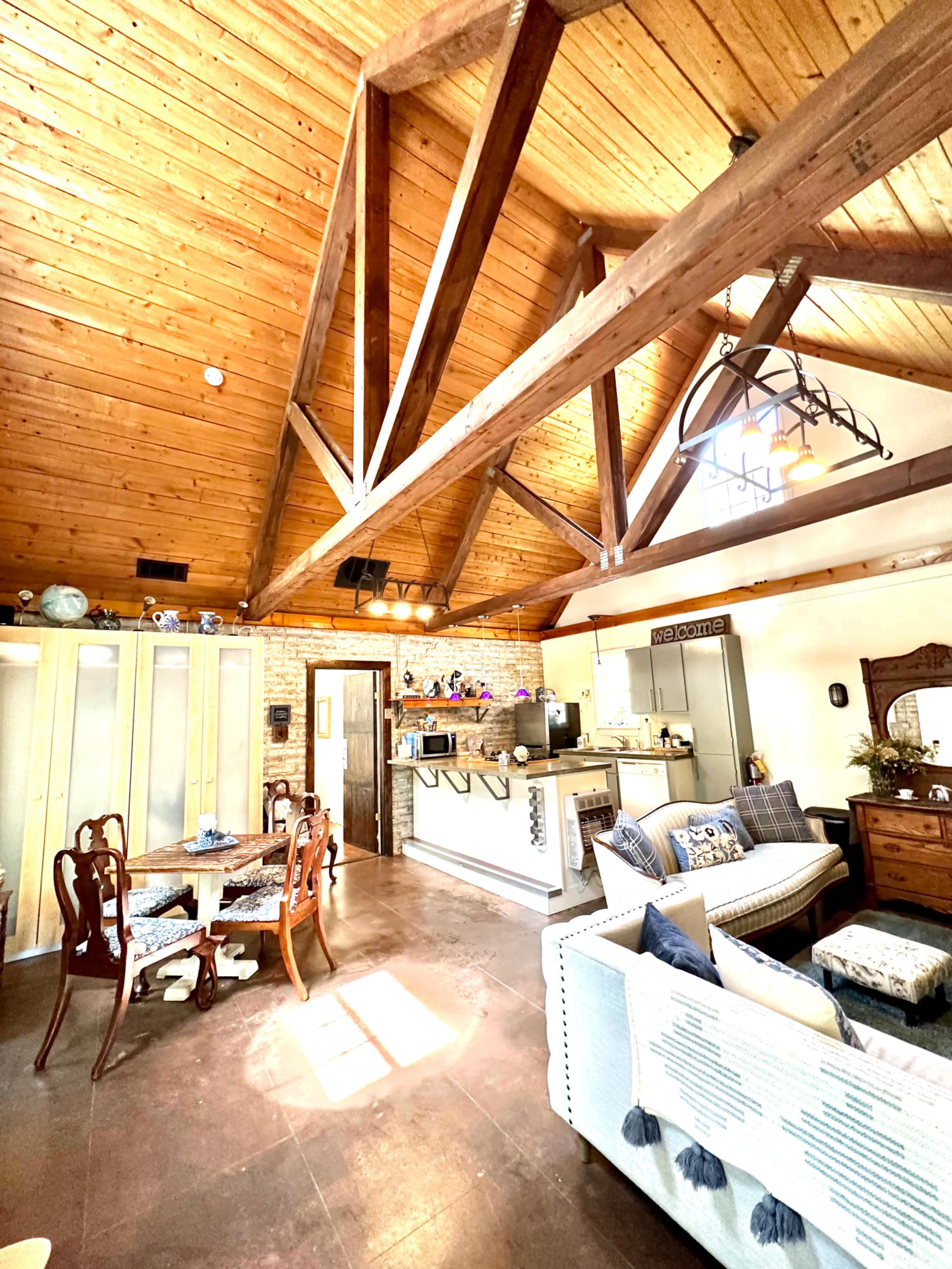 A spacious, rustic-style living area with a high wooden beam ceiling, a dining table, a sofa, and a kitchen area in the background.