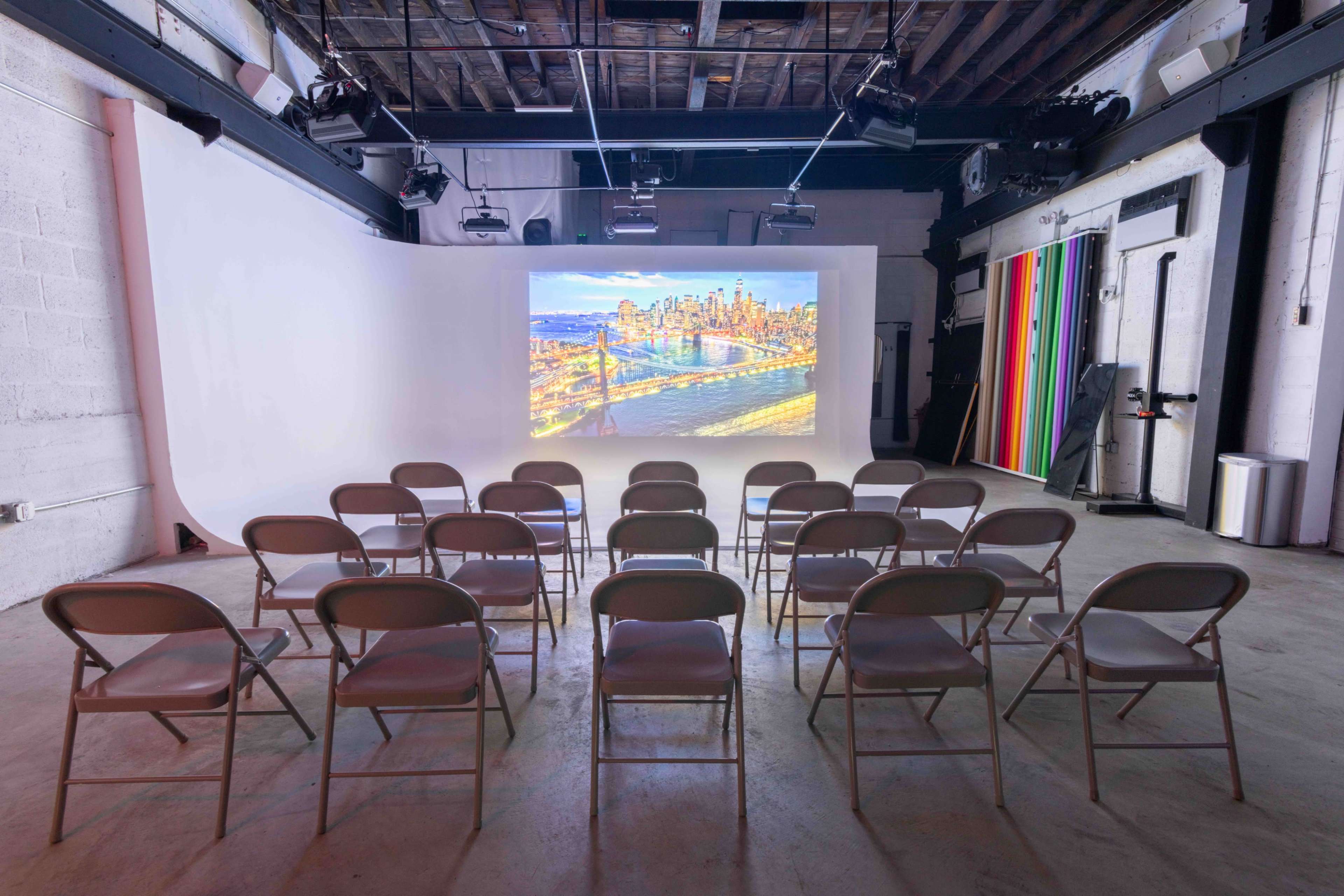 The image shows a room set up with rows of folding chairs facing a large projection screen displaying a city skyline.