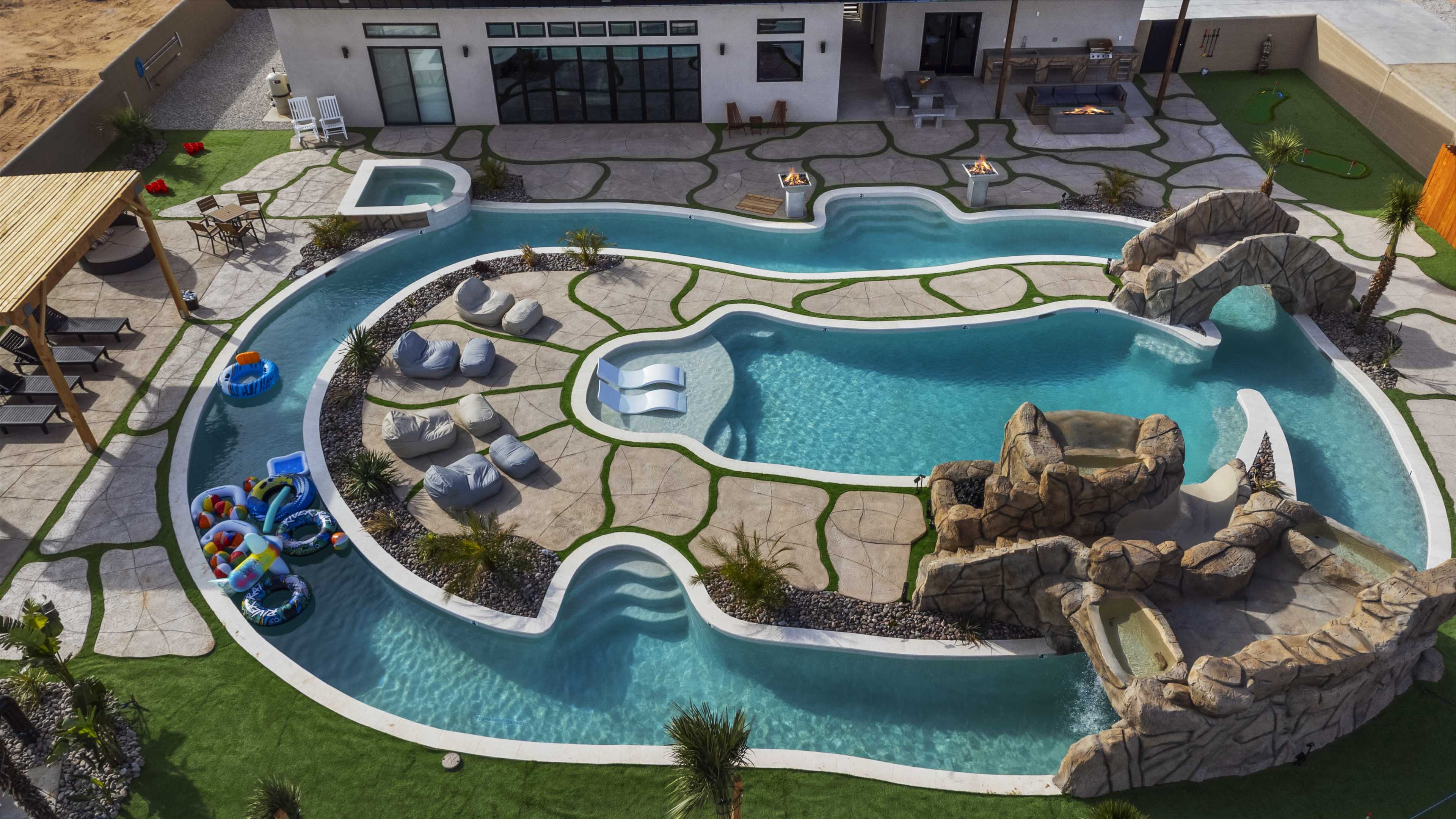 Aerial view of a large, landscaped outdoor pool with a rocky waterfall feature, surrounded by seating areas and colorful inflatable pool floats.