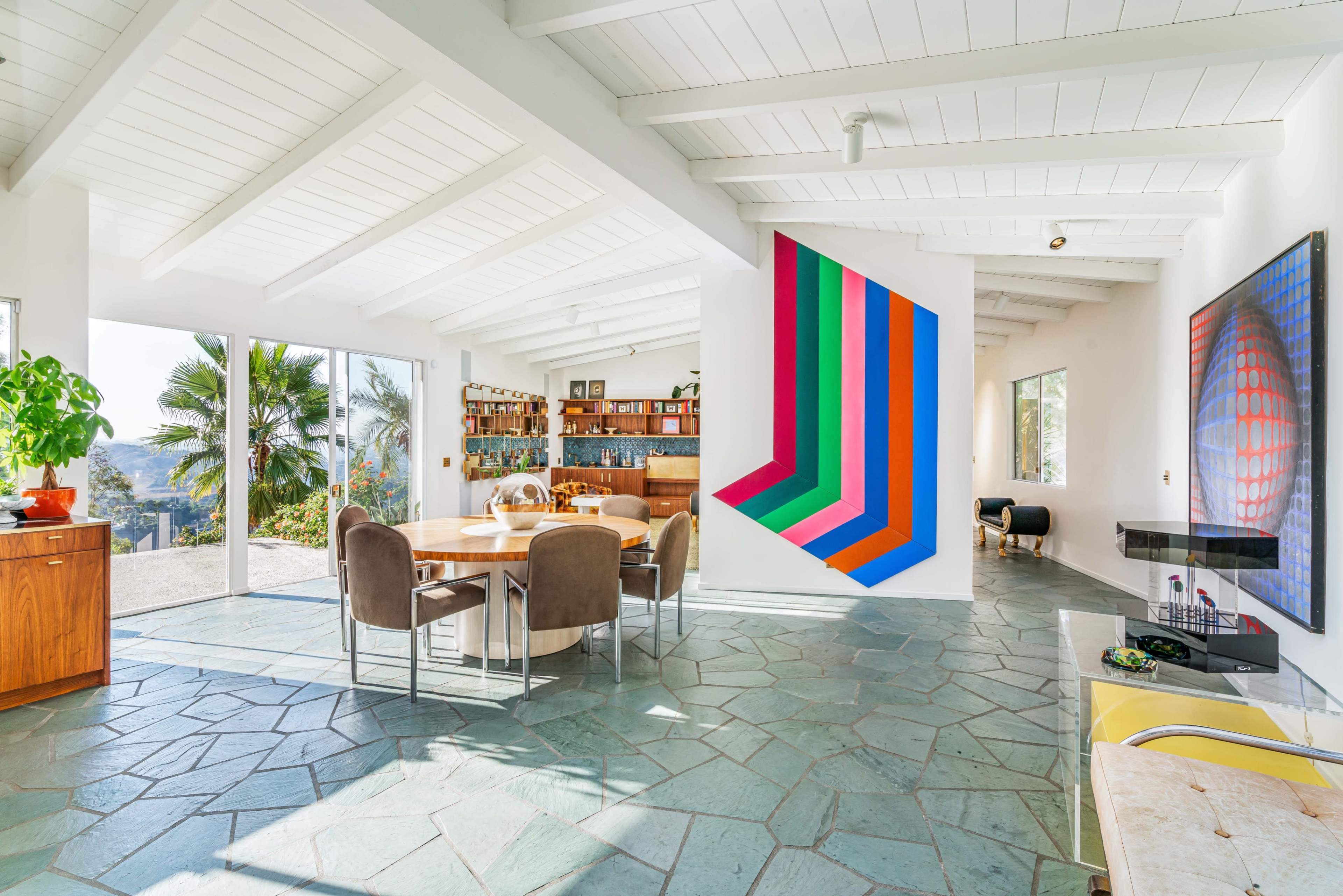 A spacious, well-lit living area with a round dining table, modern art on the walls, and a large window overlooking a palm tree and outdoor space.