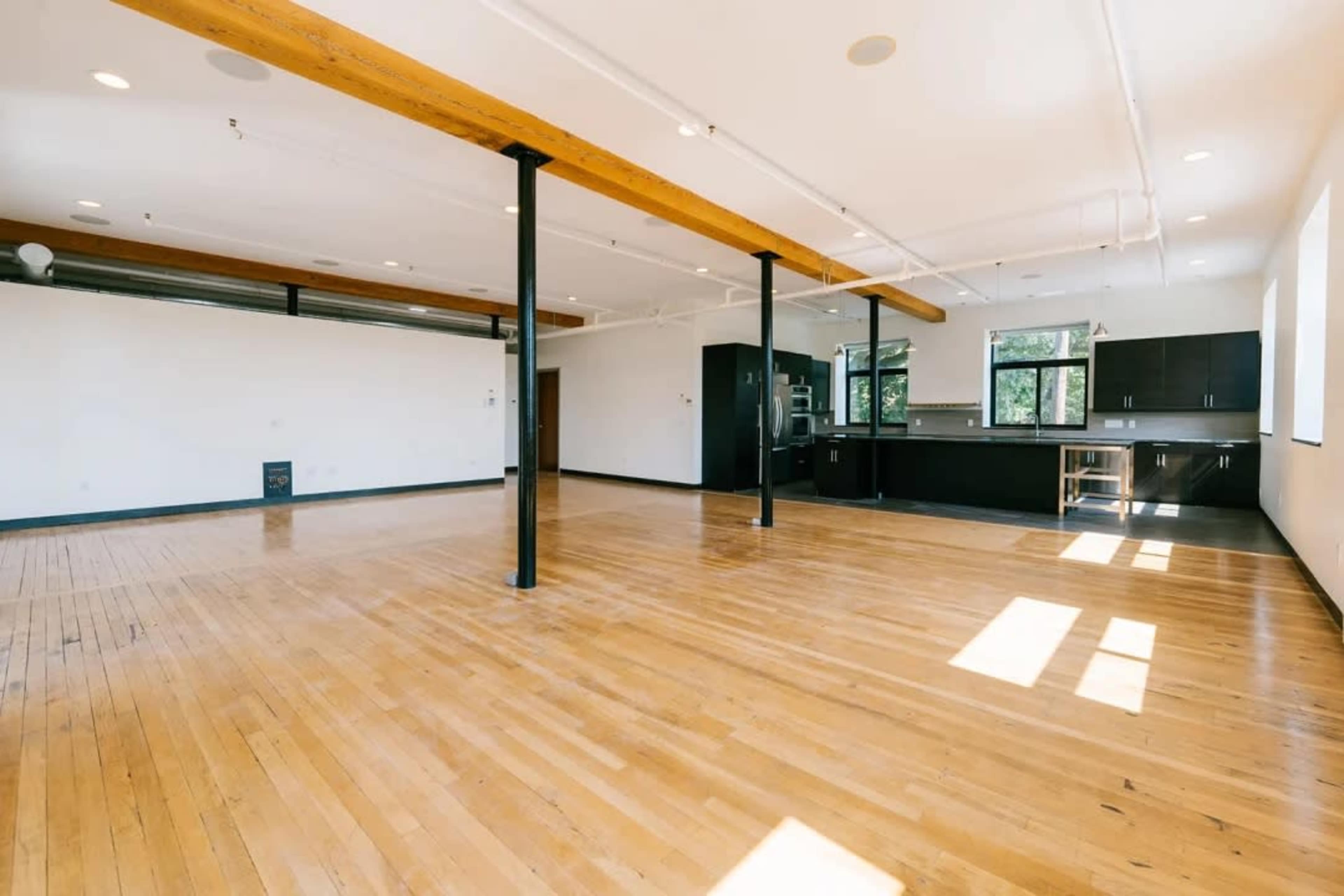 The image shows a spacious, open-concept room with hardwood floors, exposed beams, and a modern kitchen area along one side.