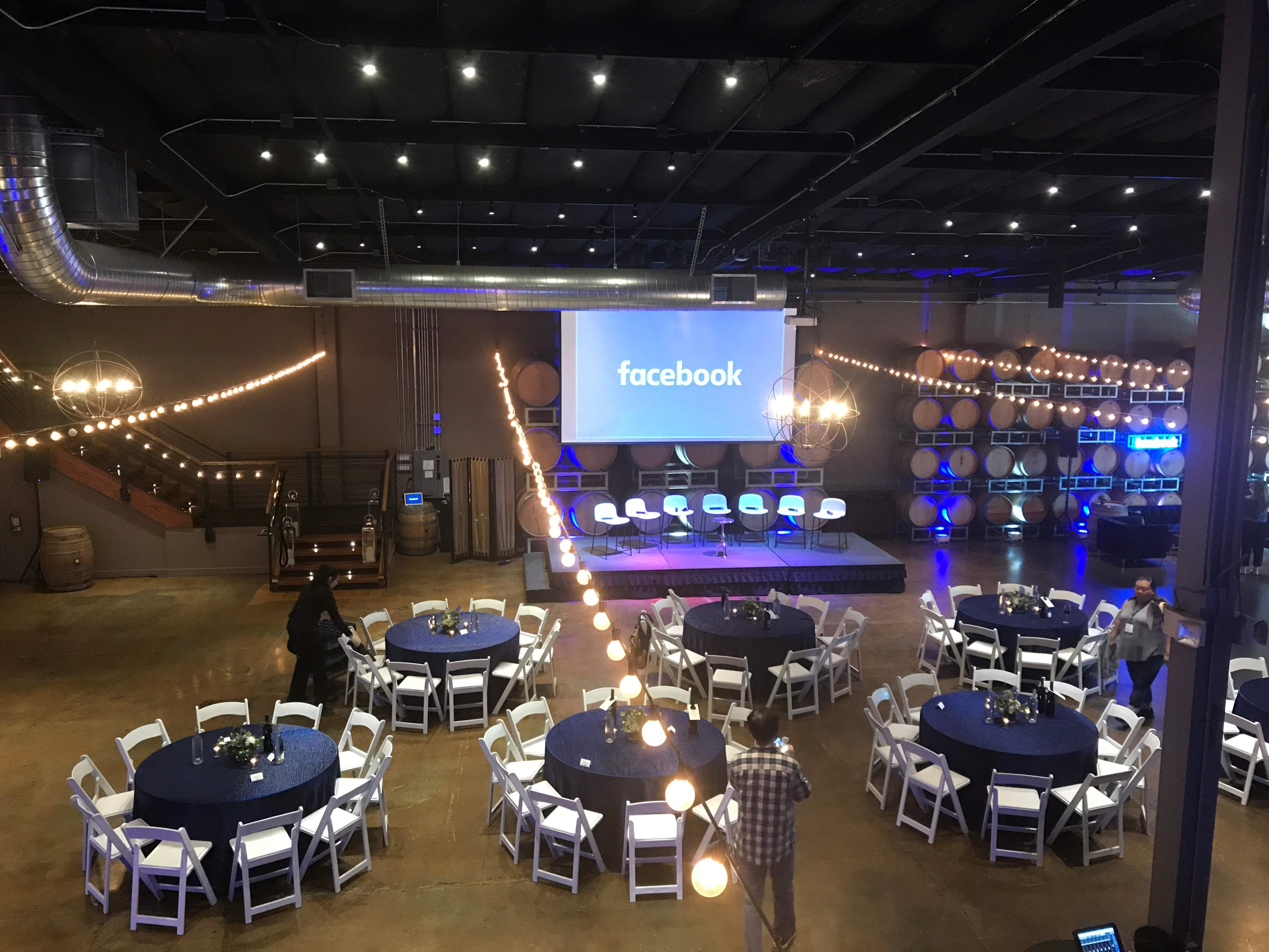 A spacious event venue features round tables set for guests, with a stage displaying the Facebook logo and decorative lighting strings overhead.