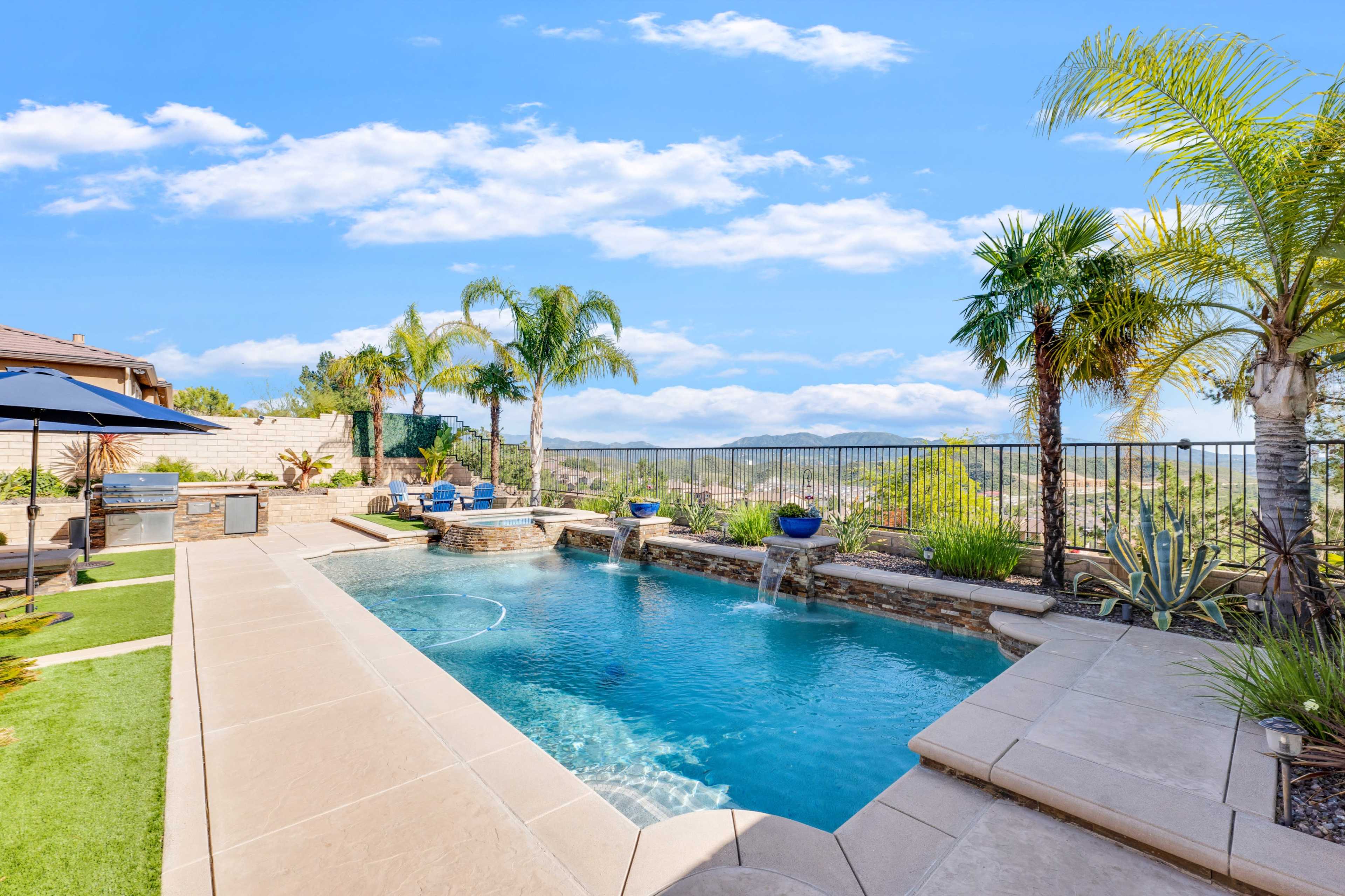 The image shows a swimming pool surrounded by palm trees and landscaped greenery, with a mountain view in the background.