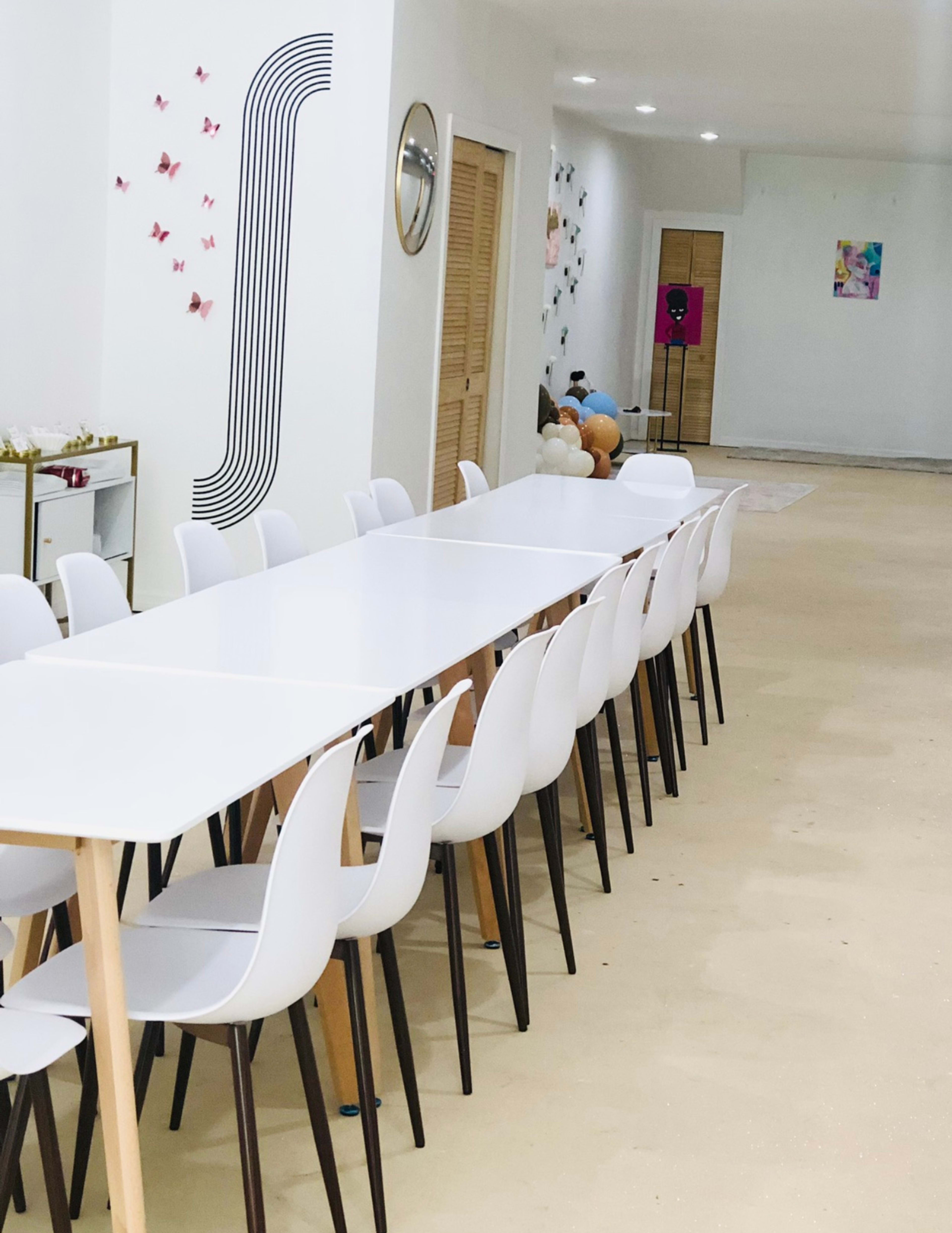 A long table with white chairs is arranged in a spacious room decorated with wall art and balloons.
