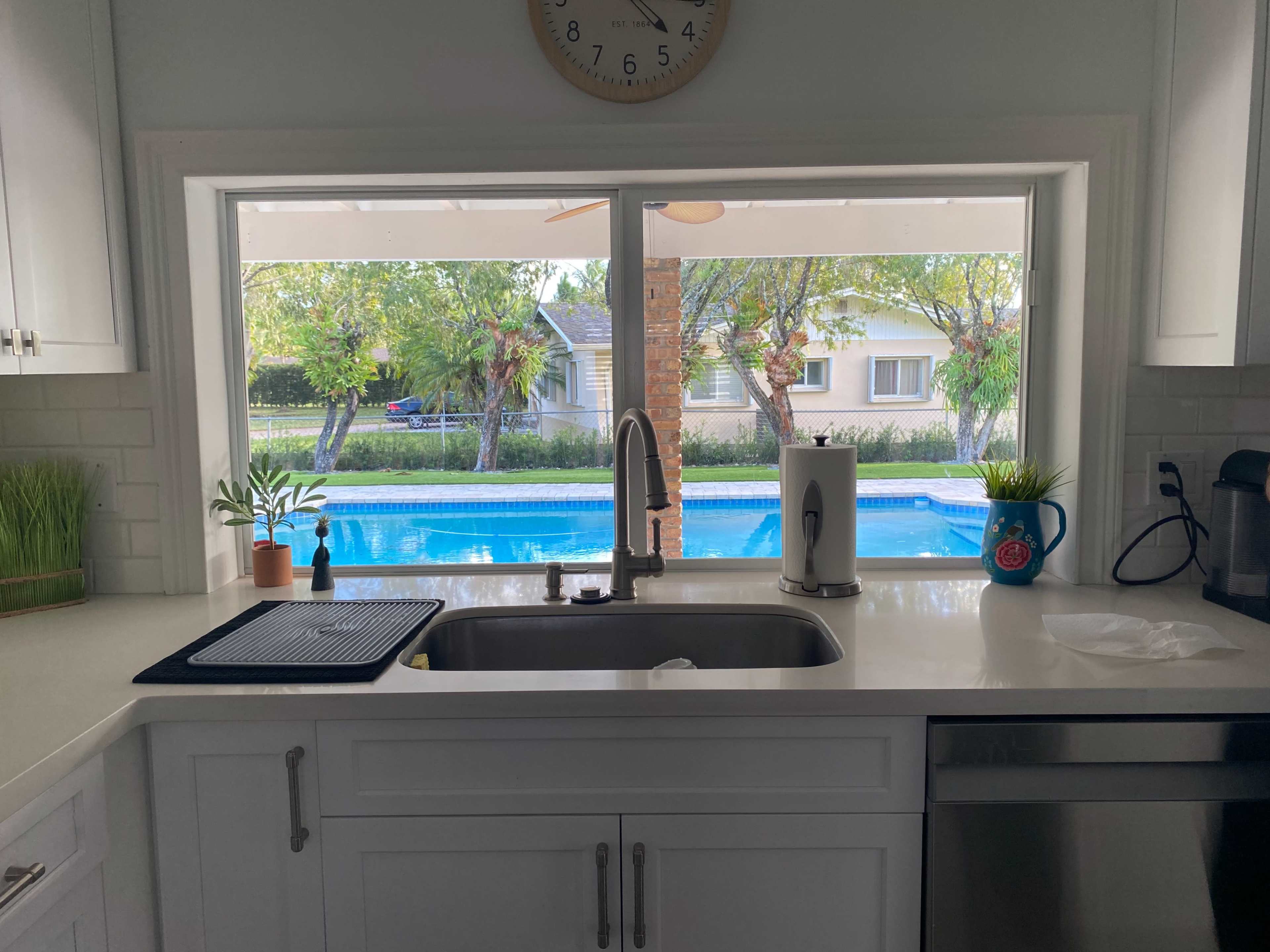 A kitchen sink is positioned in front of a window that overlooks a swimming pool and garden area.