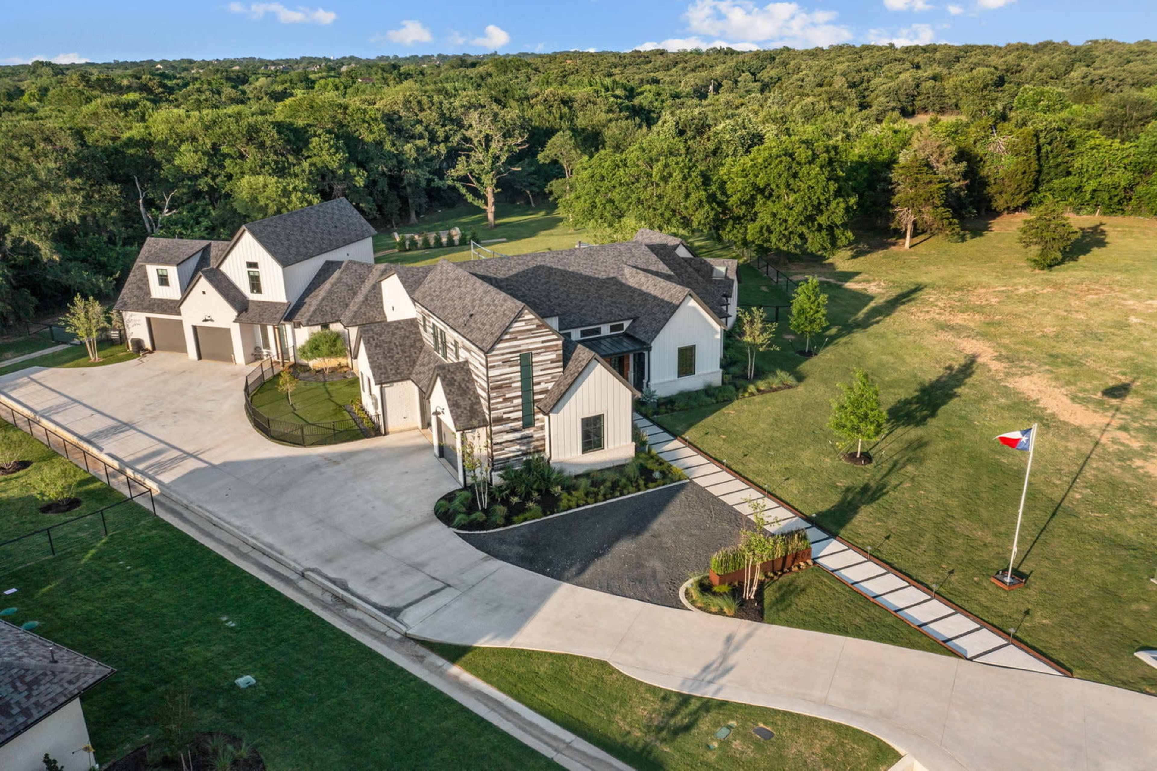 A modern, two-story house with a landscaped yard and a circular driveway is set against a backdrop of trees and green fields.