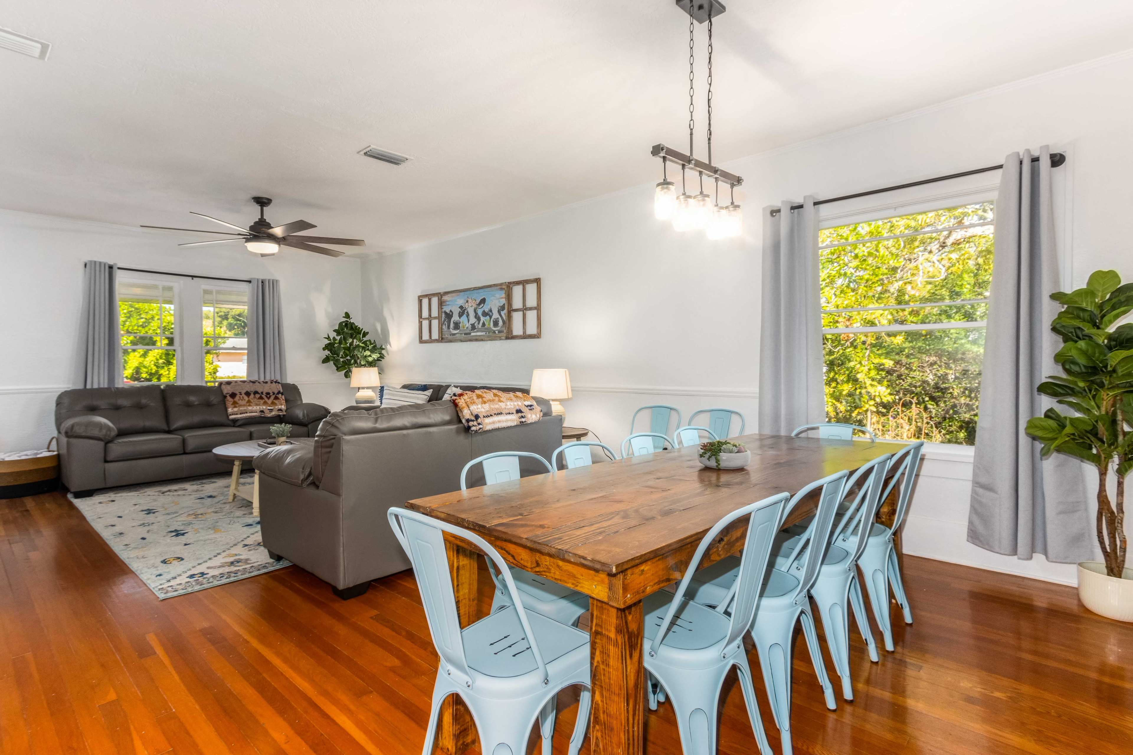 A spacious living and dining area features a wooden dining table with blue chairs, a leather couch, and large windows letting in natural light.