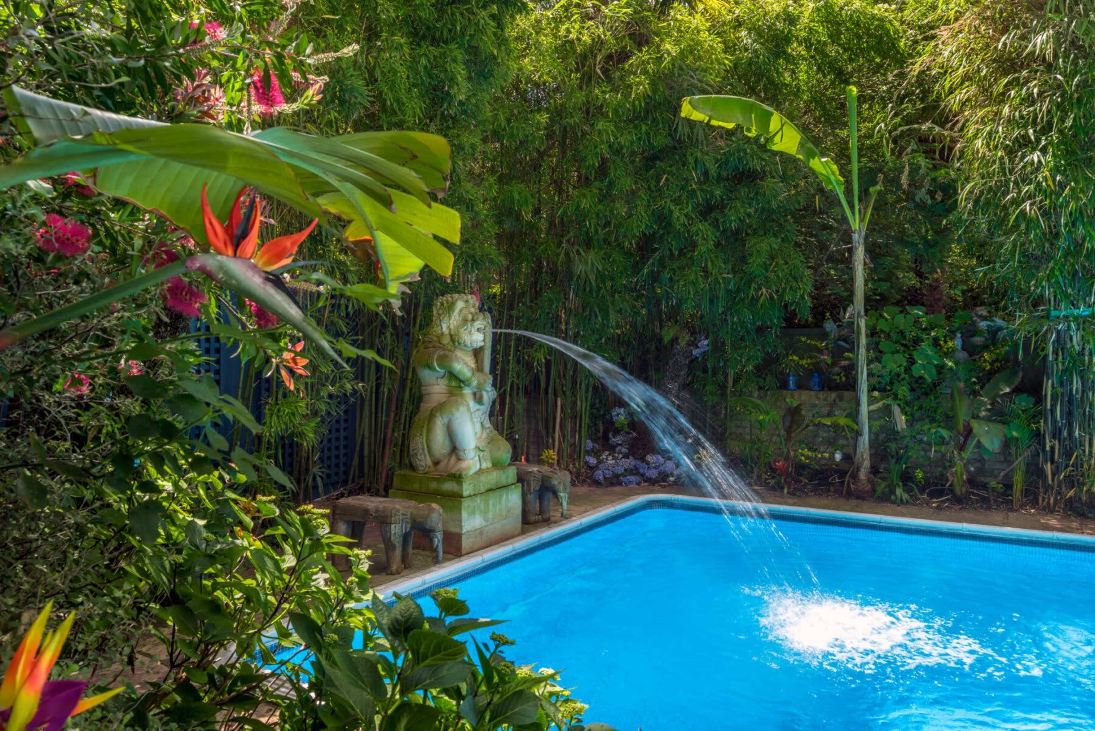 A decorative stone statue sprays water into a clear blue swimming pool surrounded by lush greenery and colorful flowers.