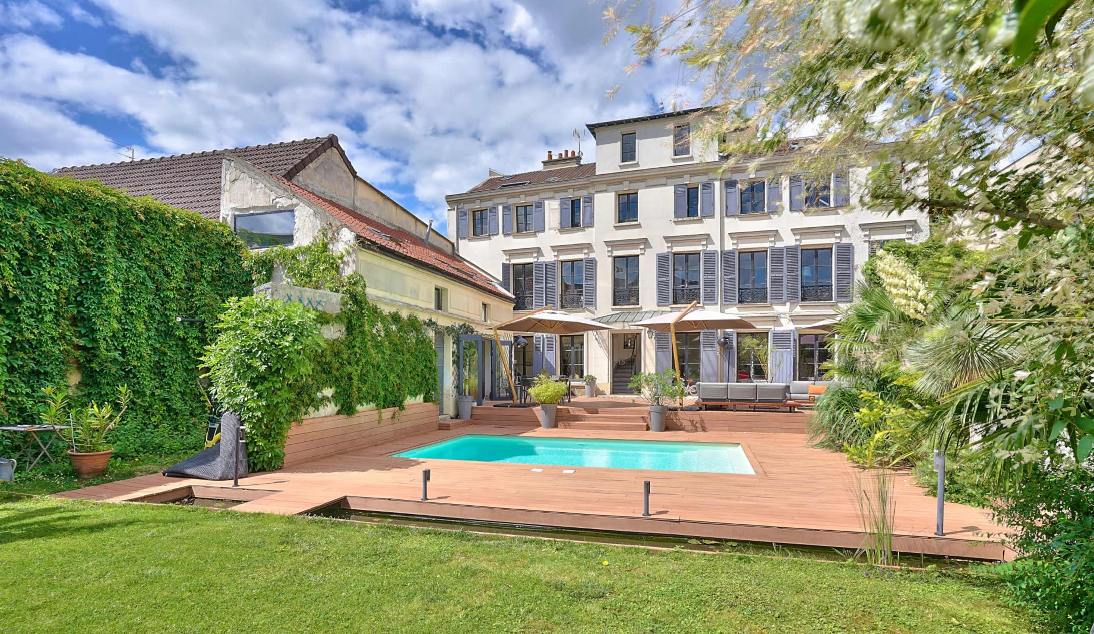A landscaped backyard featuring a swimming pool, surrounded by a wooden deck and a two-story house with multiple windows and patio umbrellas.