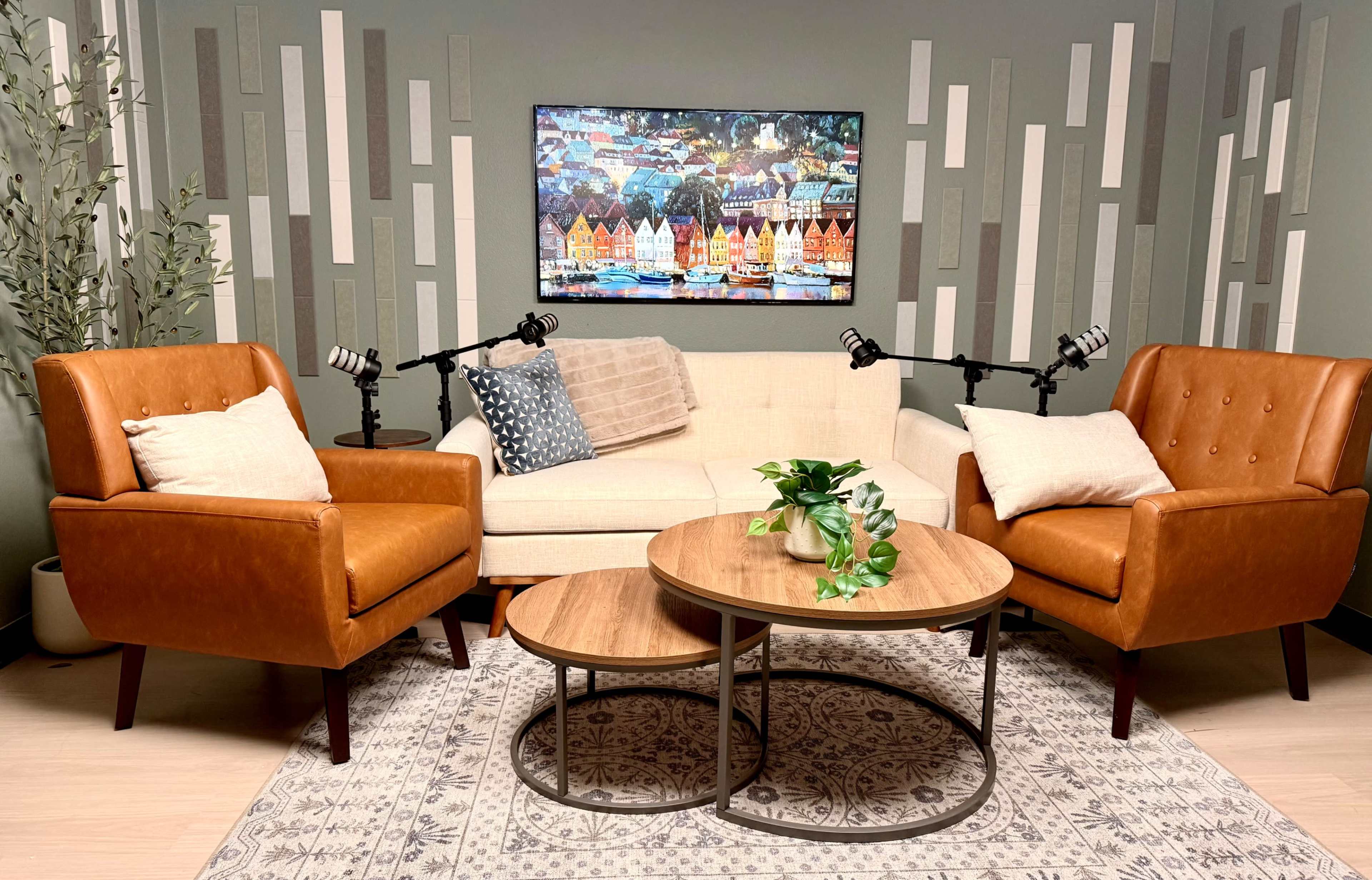The image shows a cozy living space featuring two brown leather chairs, a light-colored sofa, a round wooden table, and a colorful artwork on the wall, with two microphones positioned for a recording session.