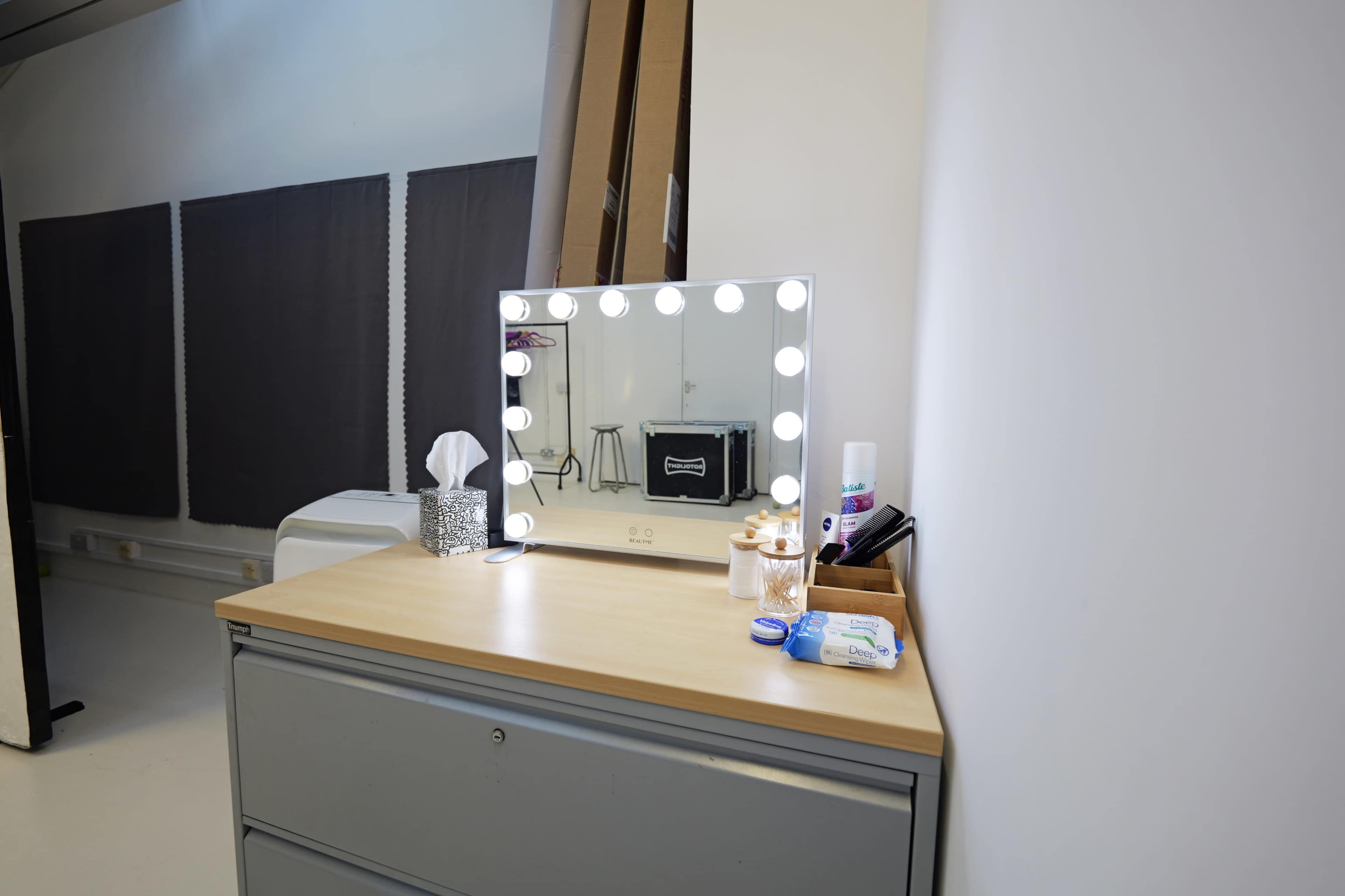 The image shows a vanity station with a lighted mirror on a wooden surface, accompanied by various items including tissues and personal care products, set against a plain wall in a room.