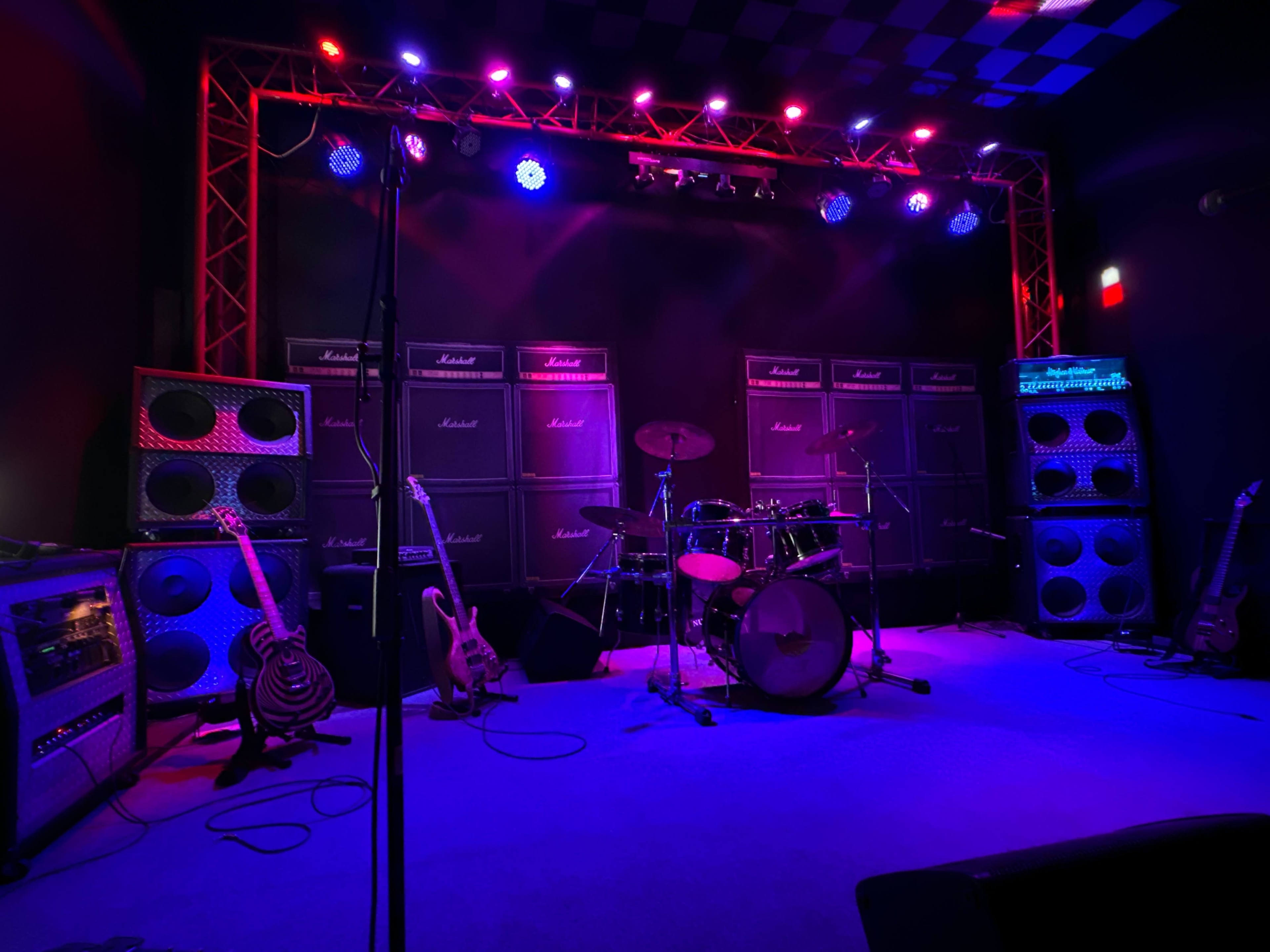 The image shows a stage setup with multiple Marshall speaker cabinets, a drum kit, and electric guitars arranged around a dark, illuminated performance area.
