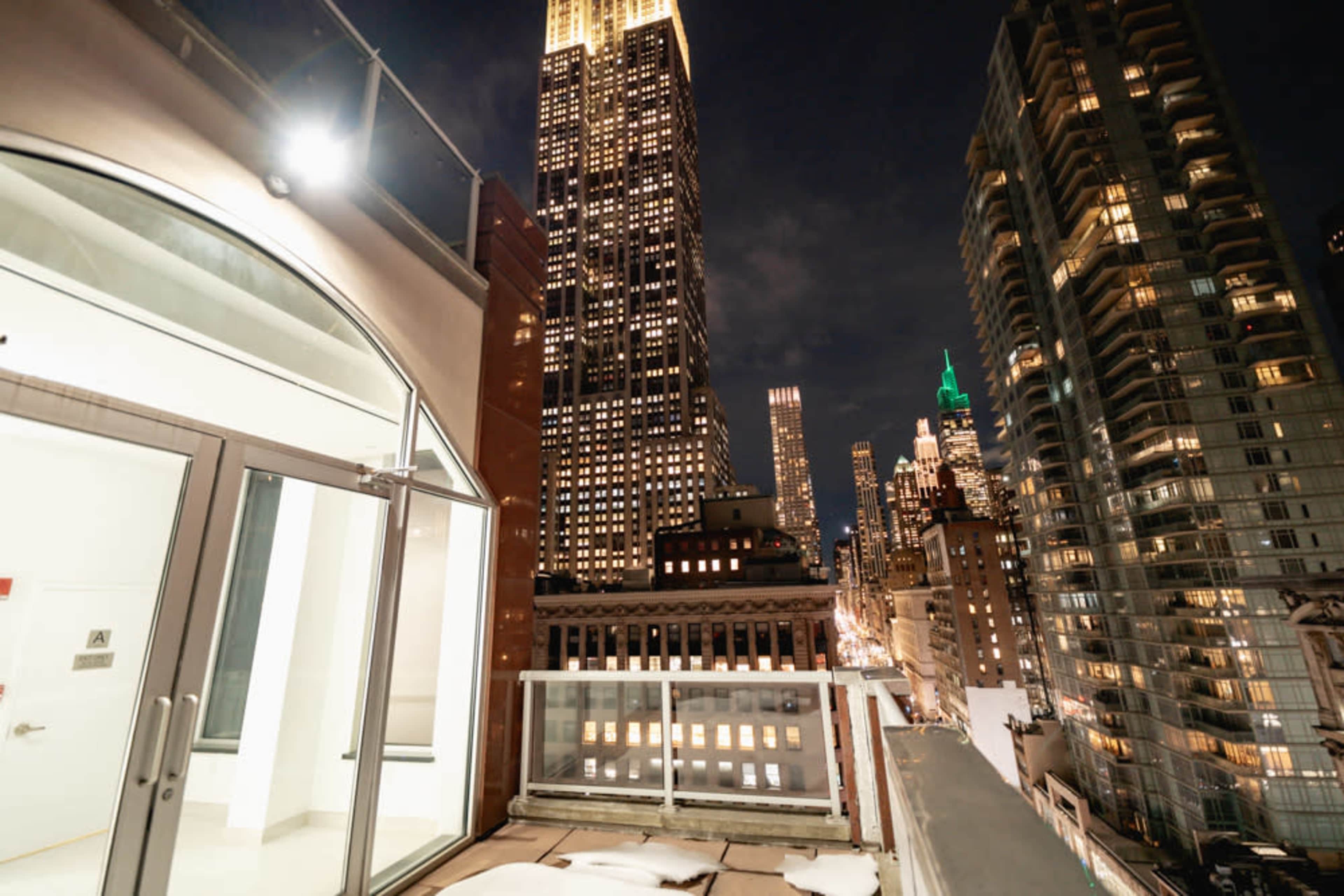 The image shows a rooftop view of a city skyline at night, featuring tall buildings and the illuminated Empire State Building.