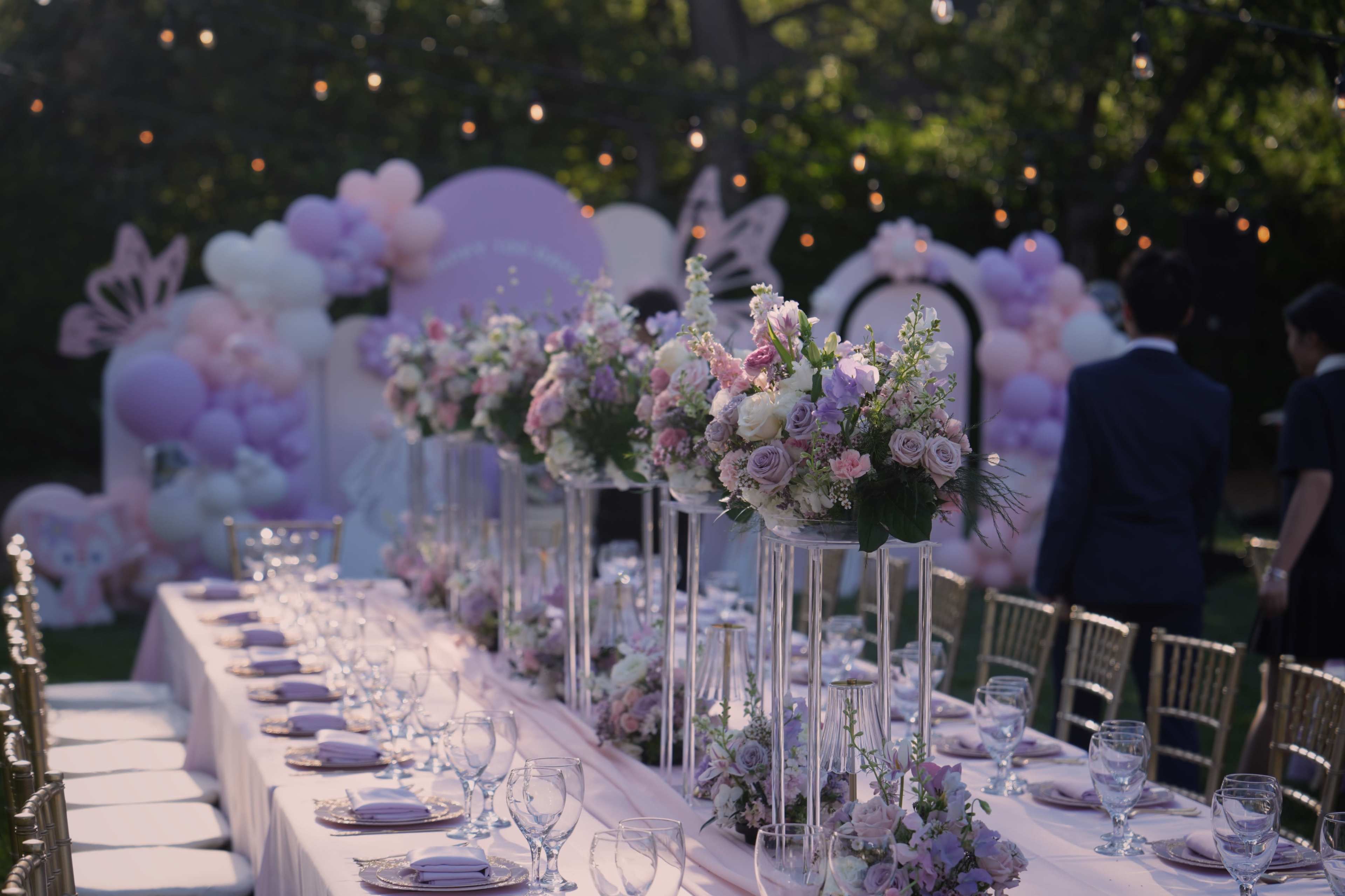 A long outdoor banquet table is set with elegant glassware and floral centerpieces, surrounded by decorations of pastel balloons and lights.