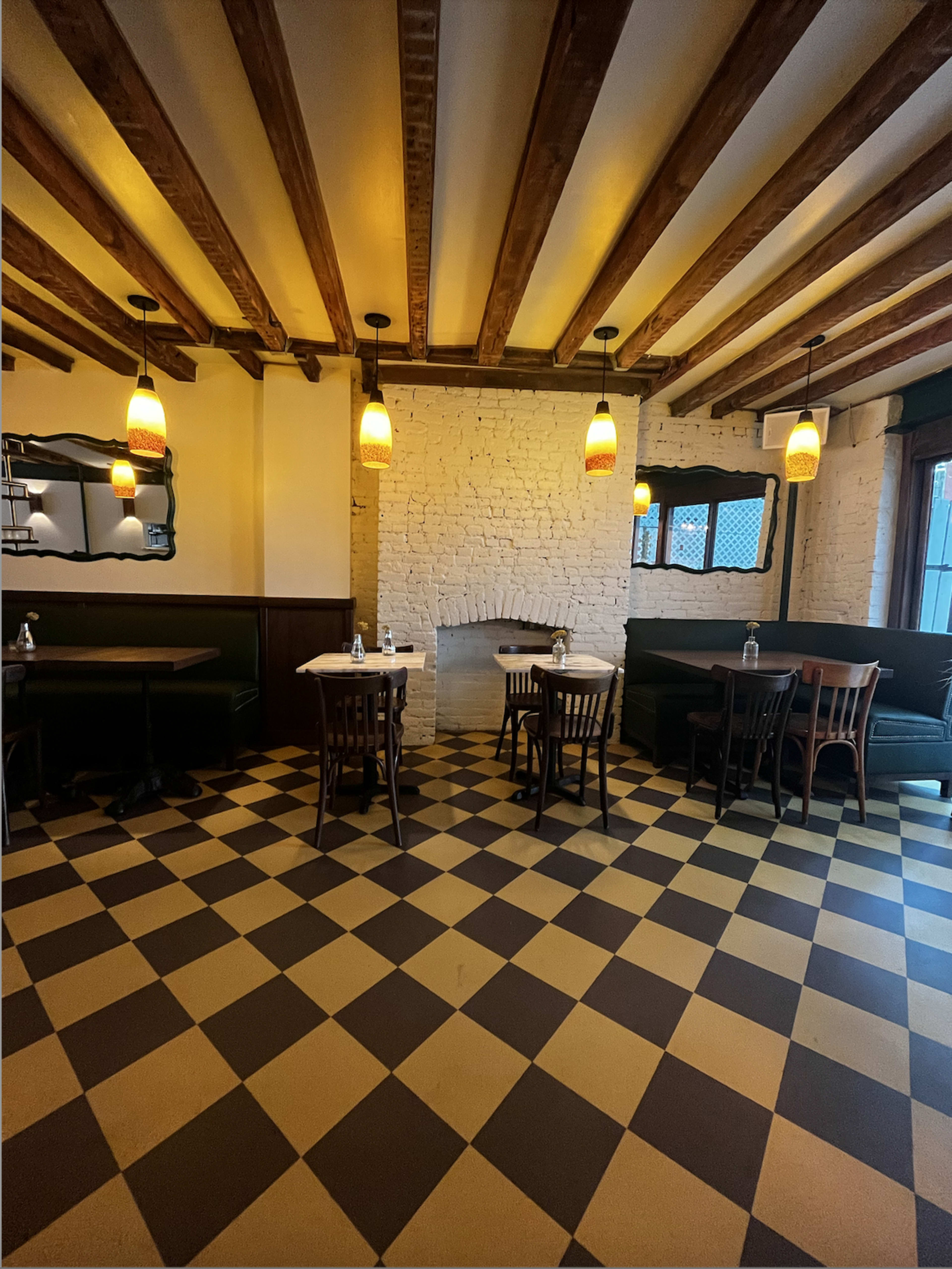 The image shows a restaurant interior with a checkerboard floor, wooden beams on the ceiling, and several tables and chairs arranged around a central fireplace.
