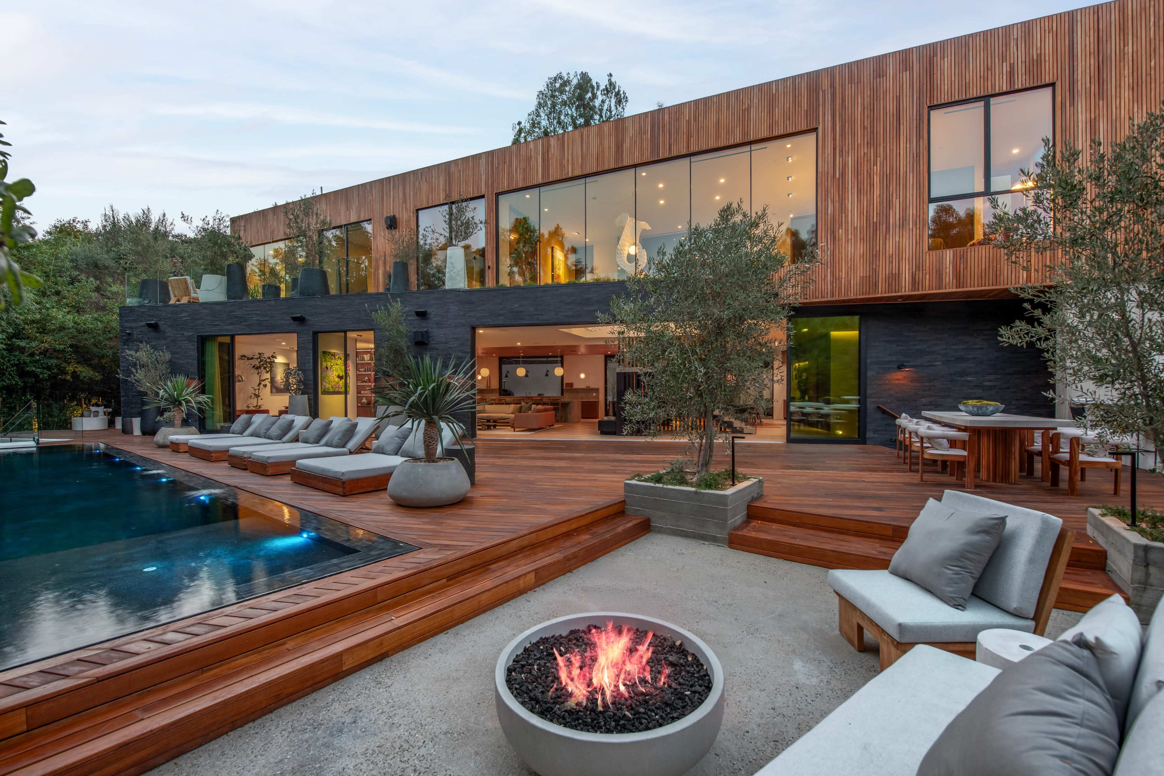 A modern two-story house with large glass windows overlooks a wooden deck and a swimming pool surrounded by lounge chairs and outdoor seating.