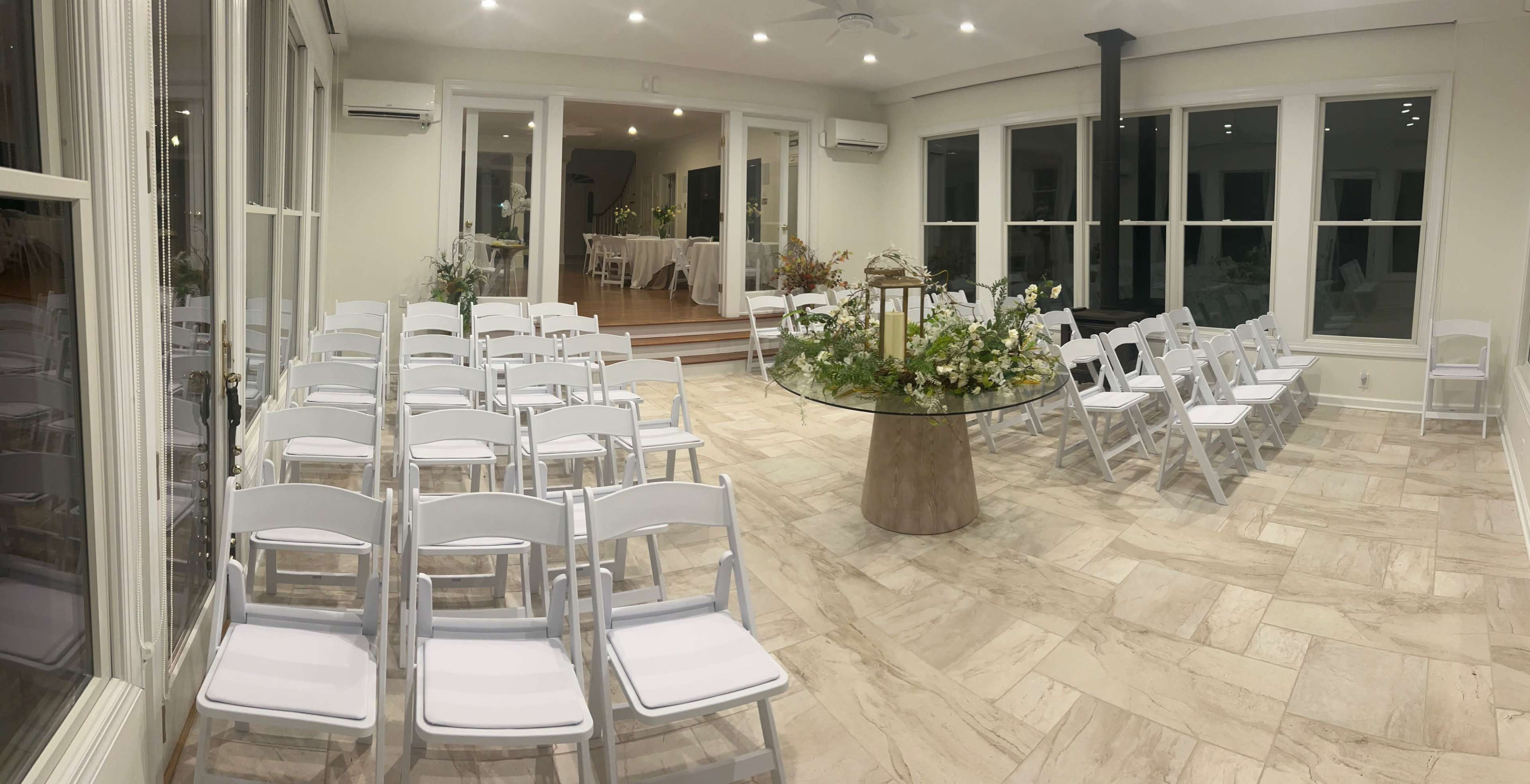The image shows a spacious indoor venue arranged with white chairs facing a round glass table adorned with floral decorations.