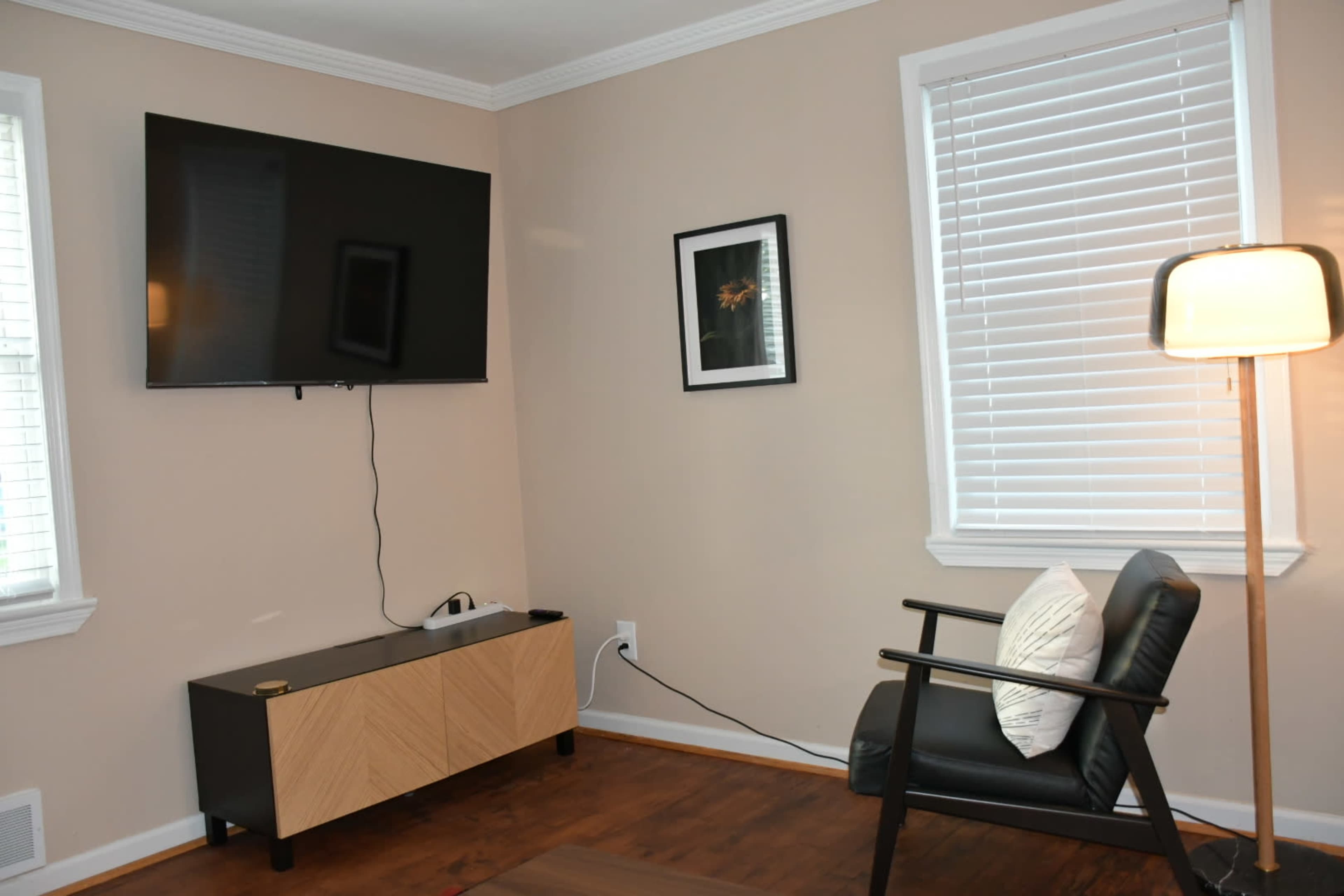 The image shows a living room corner with a mounted flat-screen TV, a small black and natural wood entertainment unit, a chair, and a lamp beside a window with blinds.
