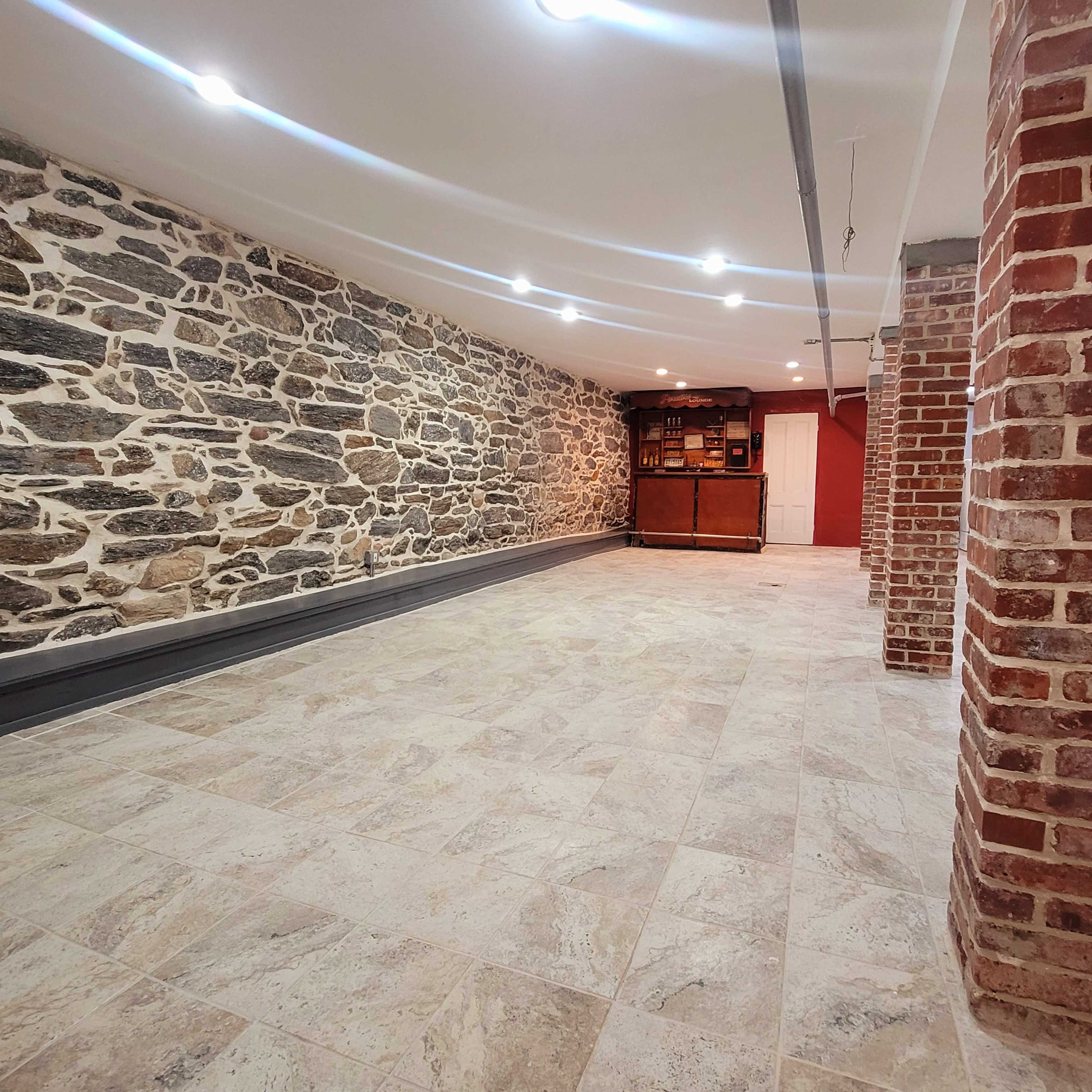 The image shows a spacious basement with a stone wall on one side, a bar setup at the far end, and brick columns lining the area.