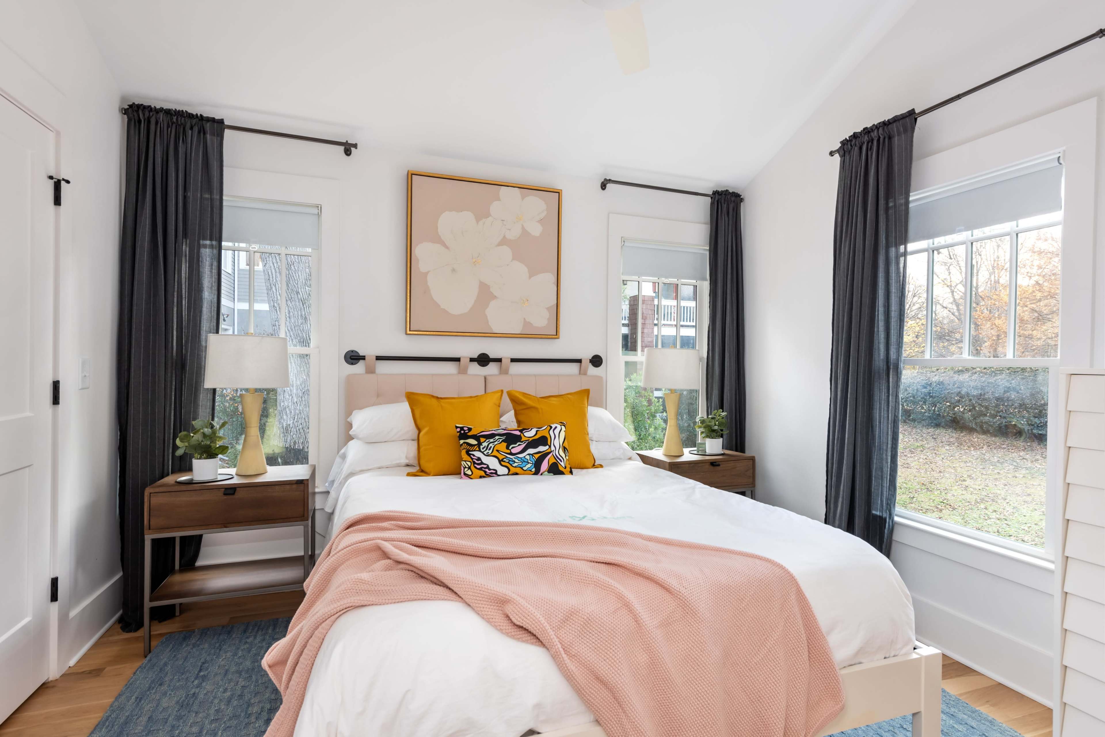 A neatly arranged bedroom features a bed with yellow and patterned pillows, flanked by wooden nightstands with lamps and plants, set against a backdrop of large windows with gray curtains.
