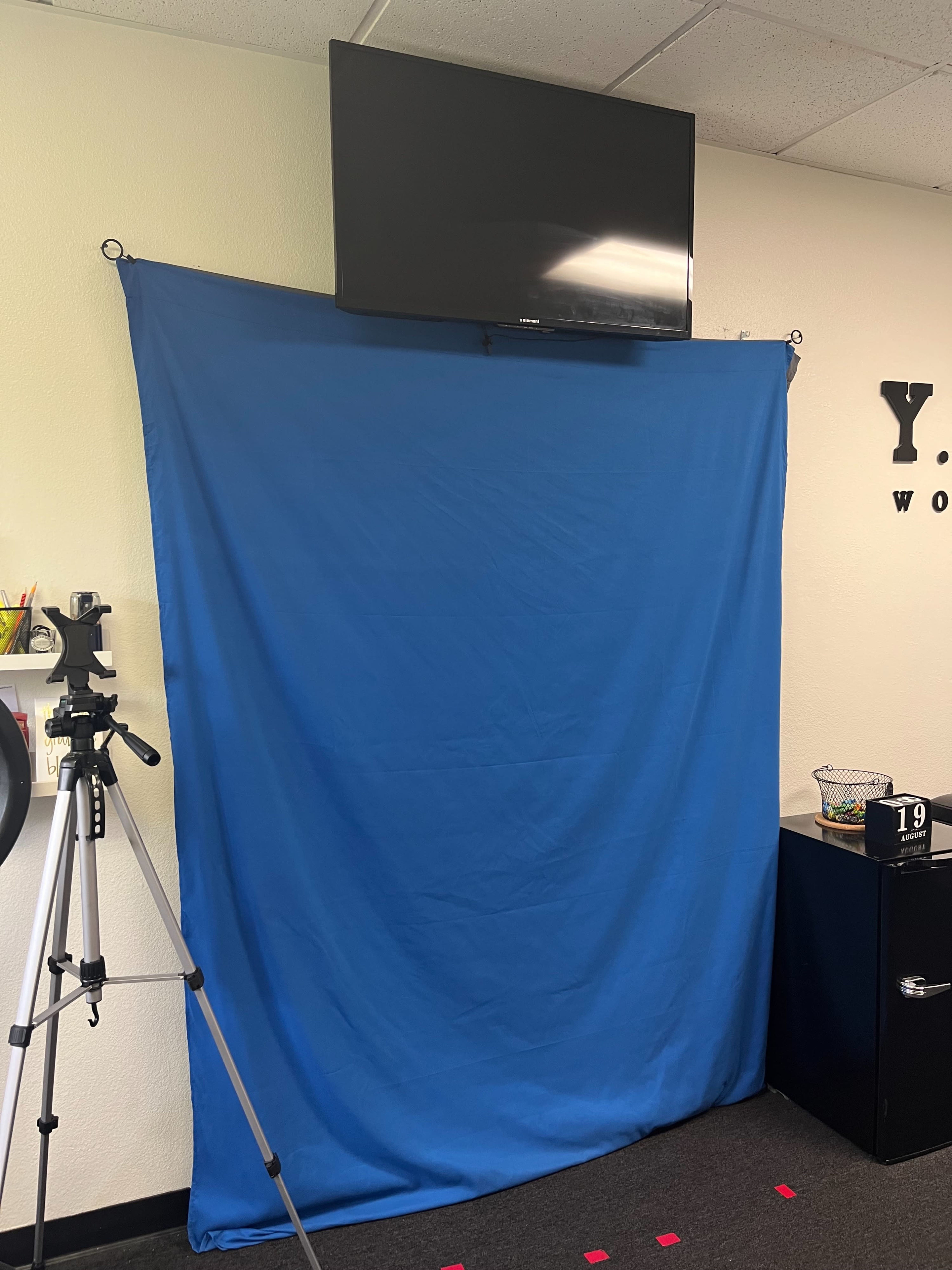 A blue backdrop hangs on the wall under a television, alongside a camera on a tripod and a black cabinet in a room.