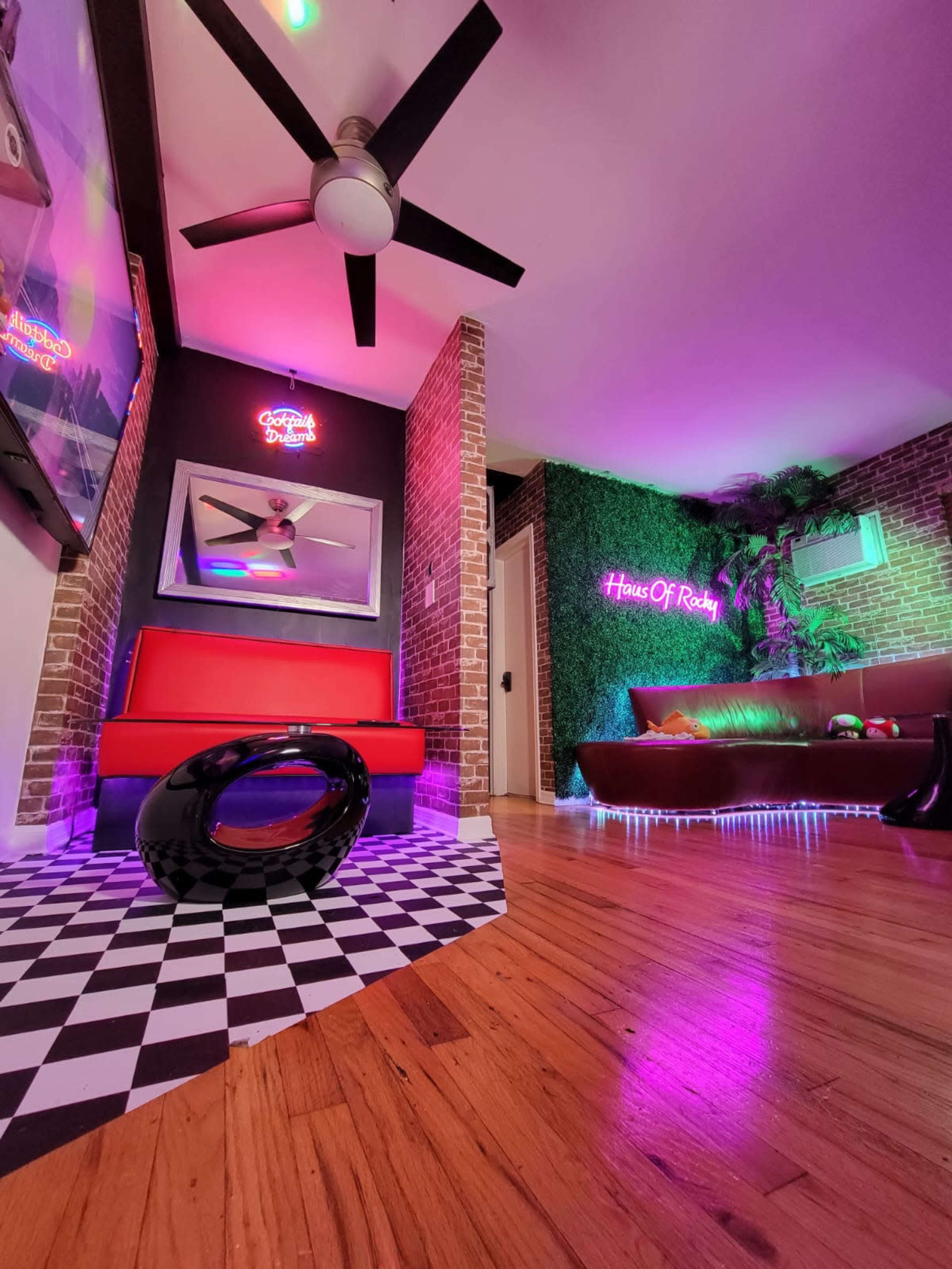 A modern room featuring a red couch, a dark brown sofa, and colorful neon lights against a brick wall and green foliage.