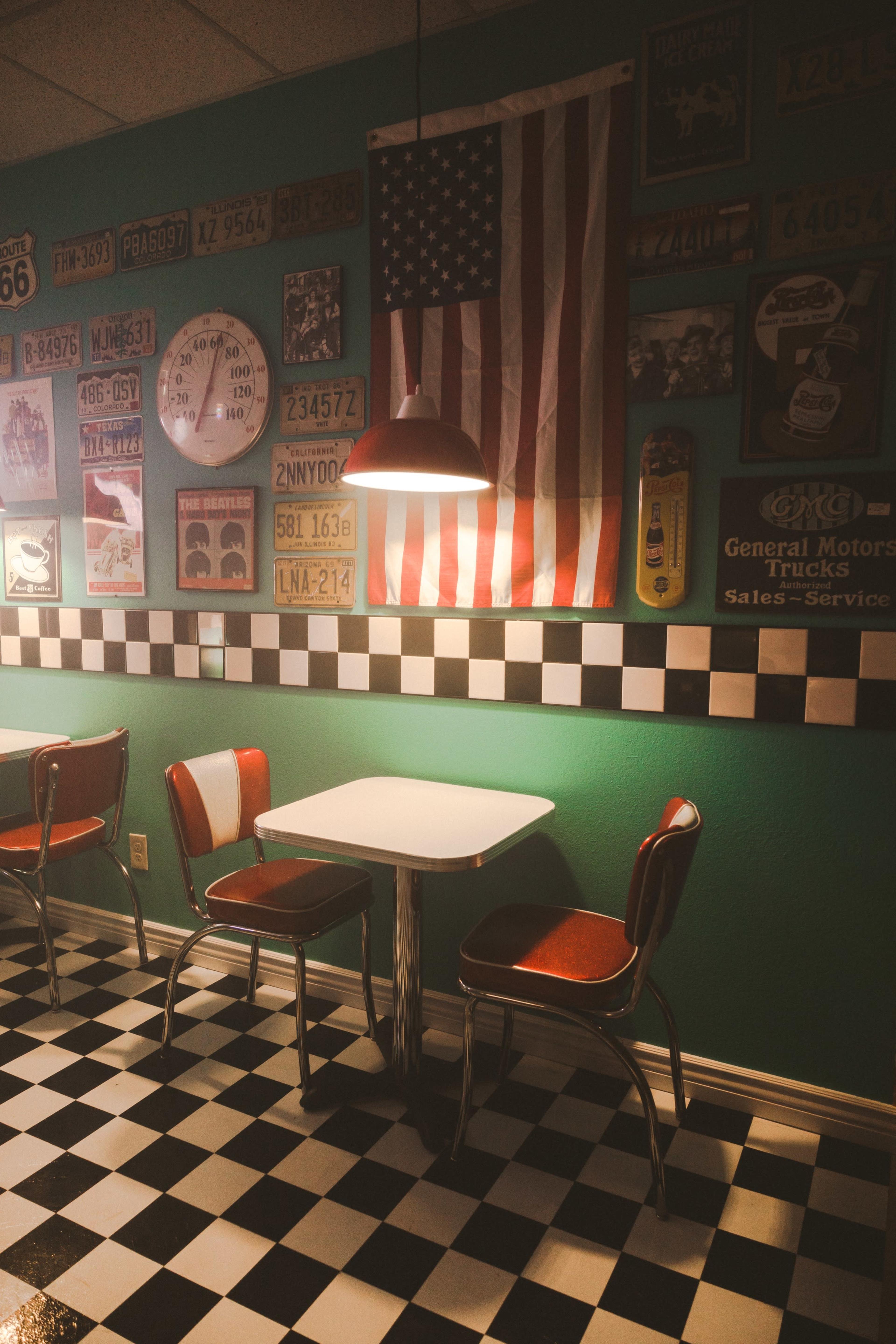 A small diner corner features a checkerboard floor, a round table with metal chairs, and vintage American memorabilia on the walls, including an American flag.
