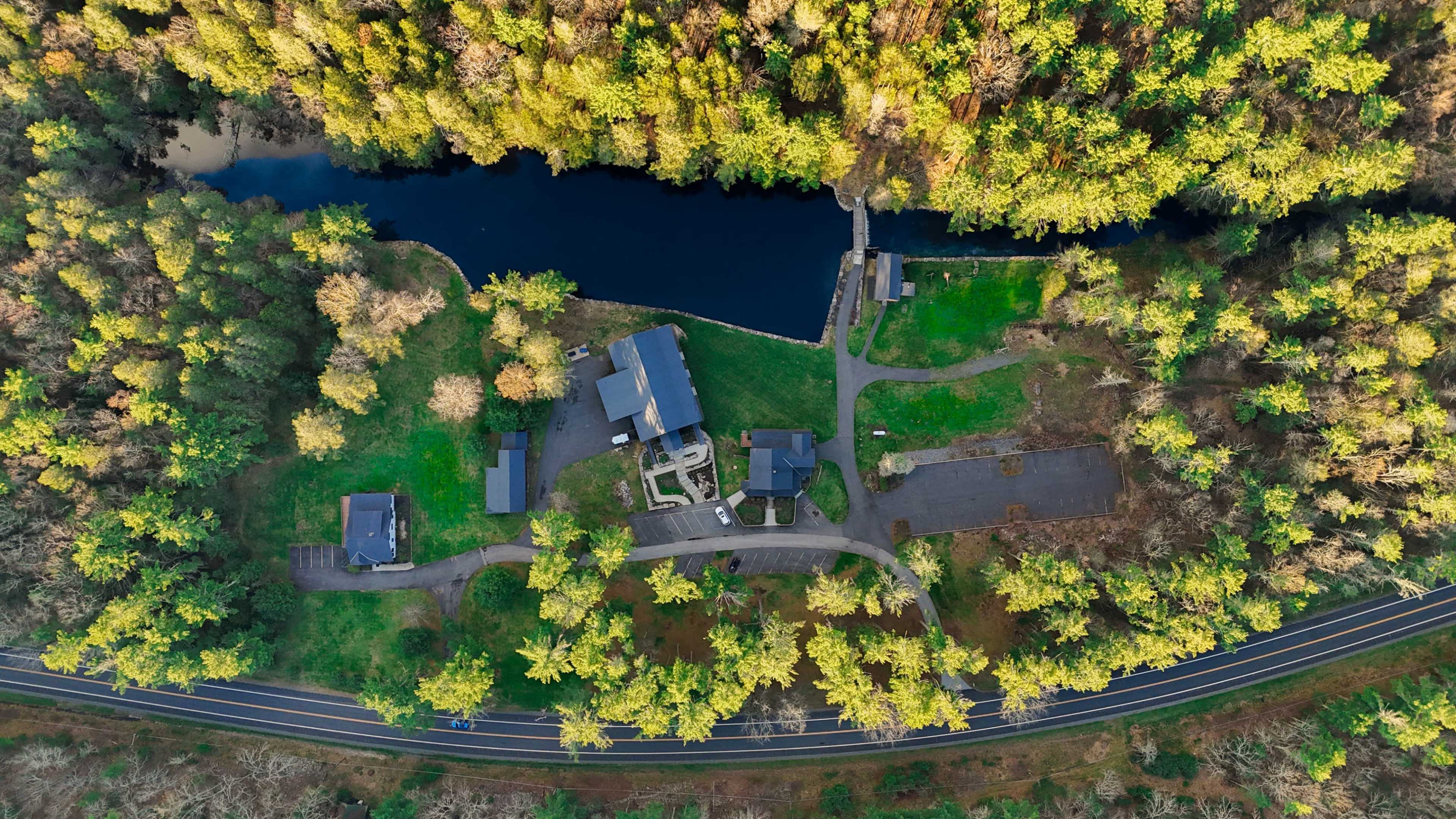 The image shows an aerial view of a large property with several buildings and green landscaping adjacent to a body of water, bordered by a road.