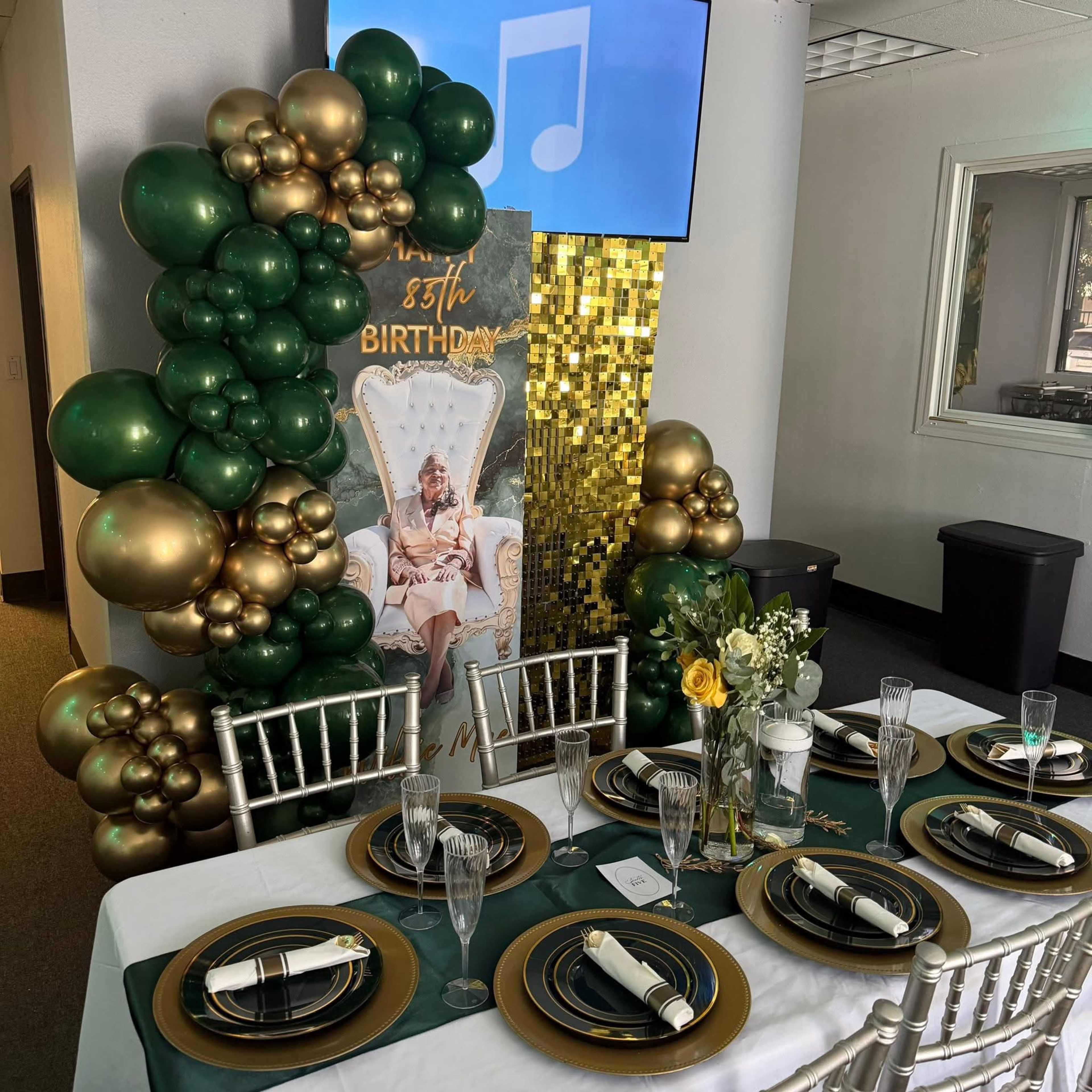 The scene features a decorated birthday party table set with gold and green balloons, elegant tableware, and a backdrop celebrating a birthday.