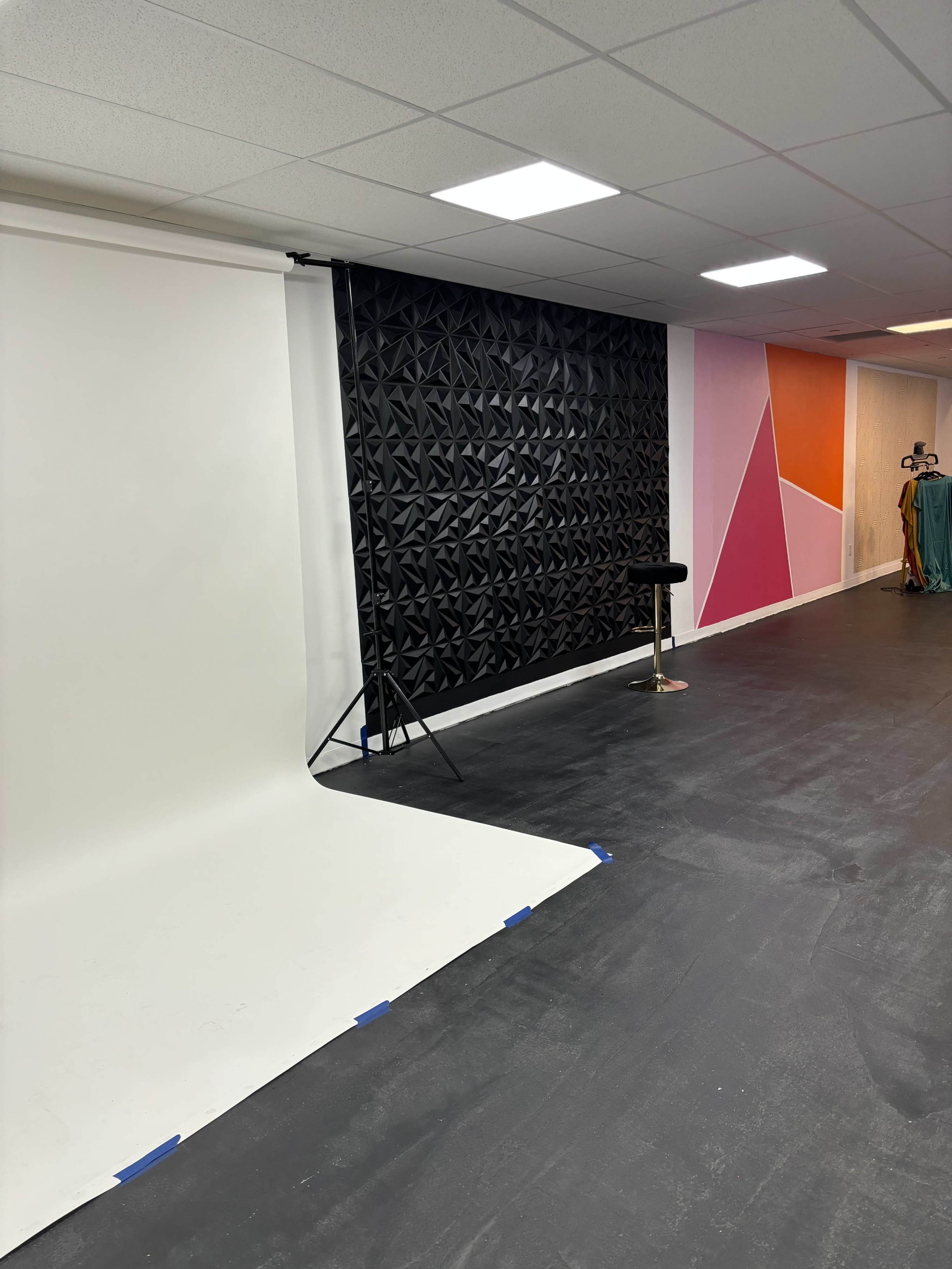 The image shows a photography studio with a white backdrop on one side and a textured black backdrop on the other, along with colorful wall sections and a clothing rack in the background.