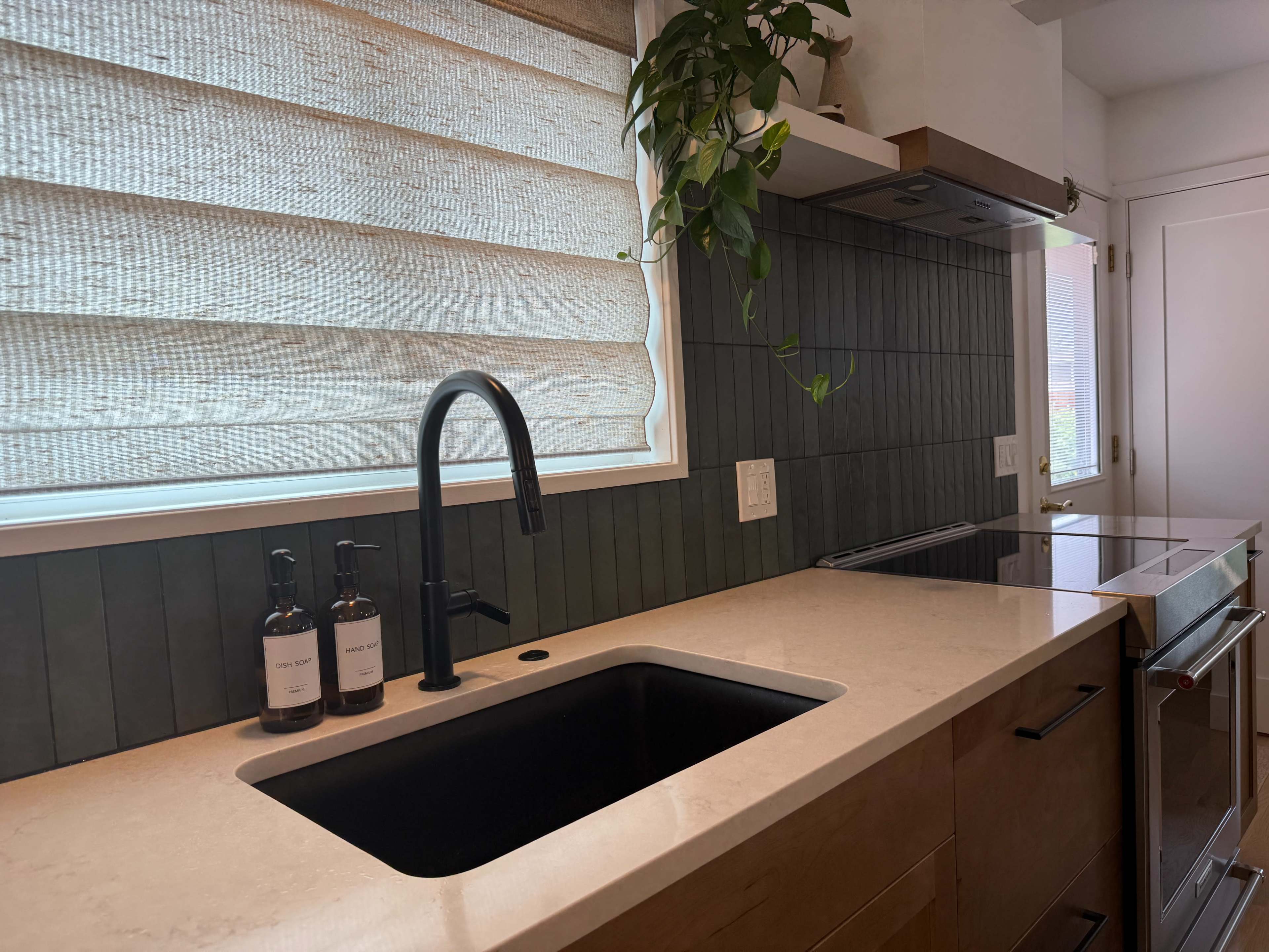 A modern kitchen features a black sink and faucet, a stone countertop, and a window with shades, accented by green plants and minimalist decor.