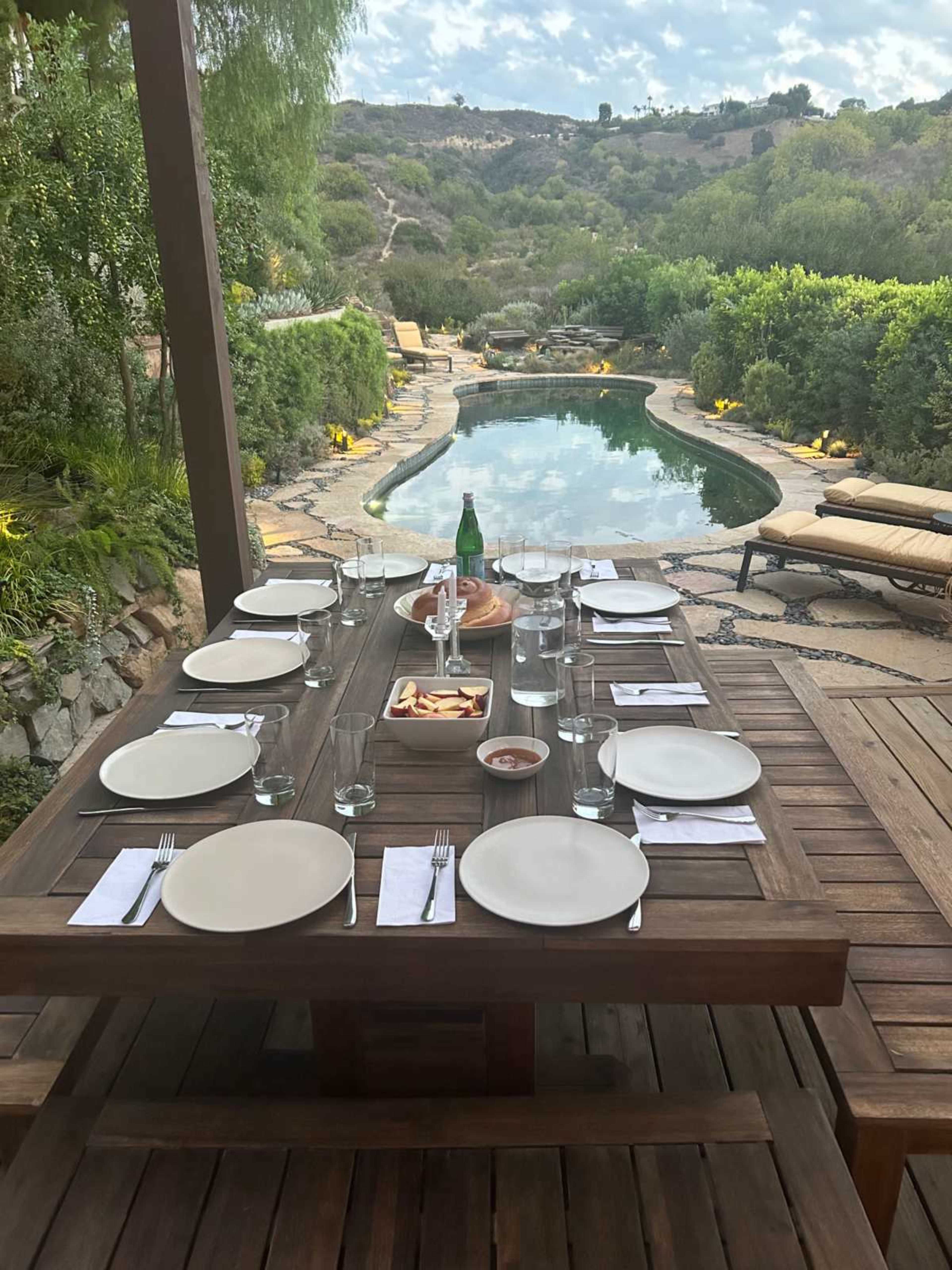 A wooden dining table is set for a meal, facing a swimming pool surrounded by lush greenery and a hillside in the background.