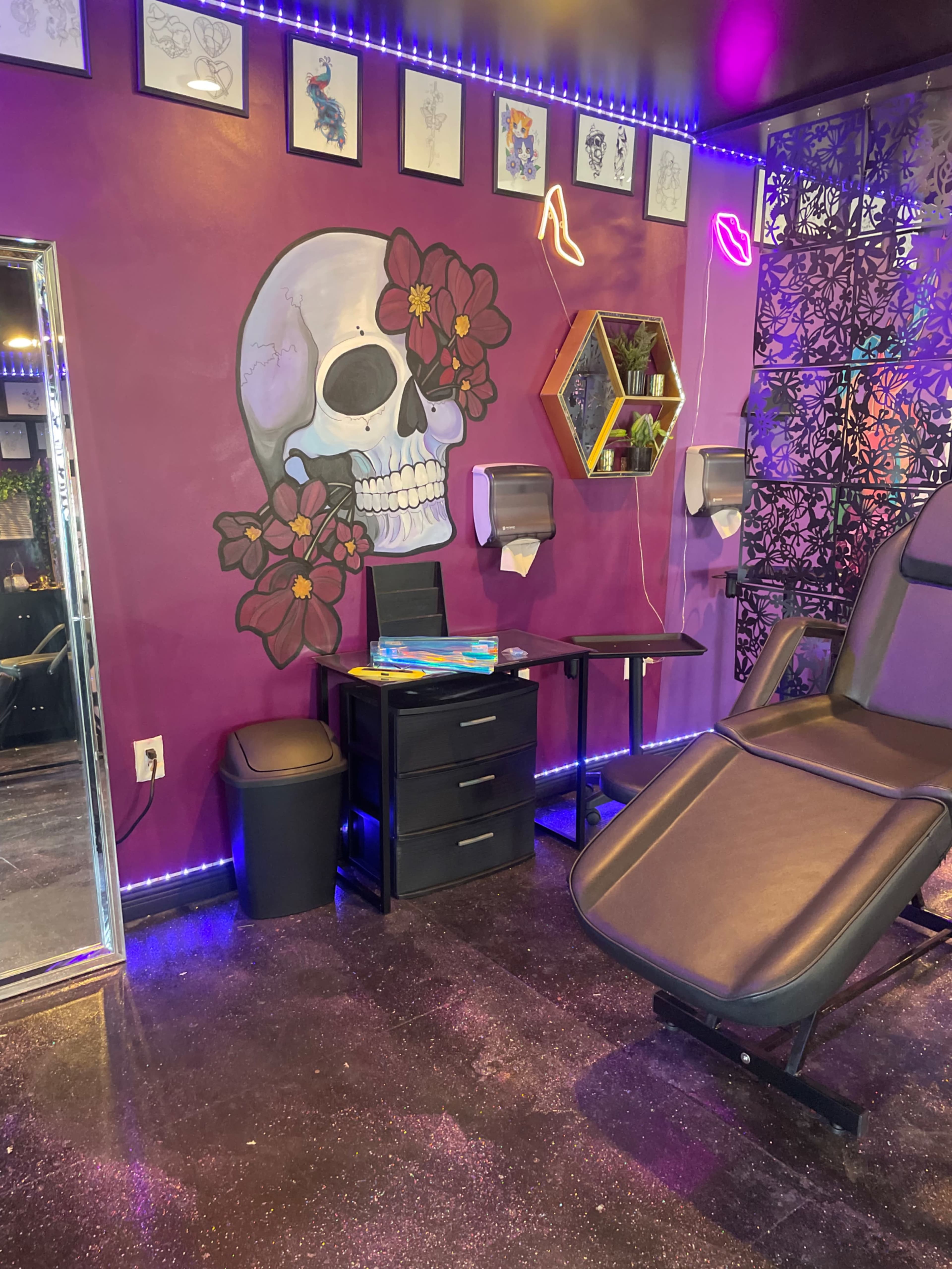 A tattoo parlor featuring a purple wall with a large skull mural adorned with flowers, a black treatment chair, and a mirrored wall with small artwork displayed above.