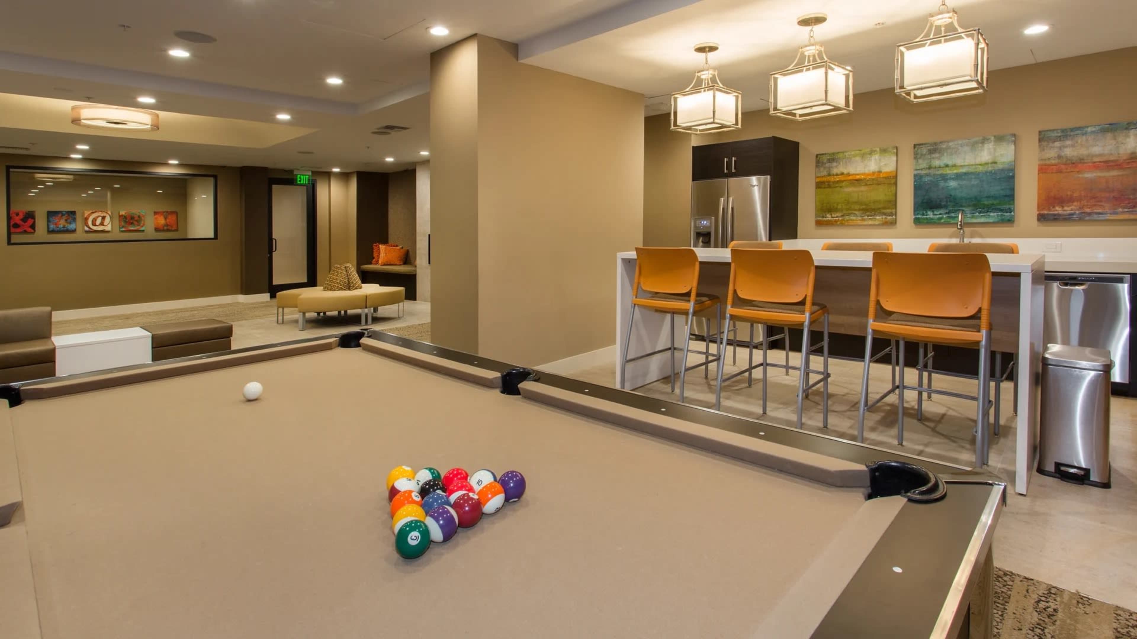 The image shows a modern lounge area featuring a billiard table, a bar with high stools, and decorative artwork on the walls.