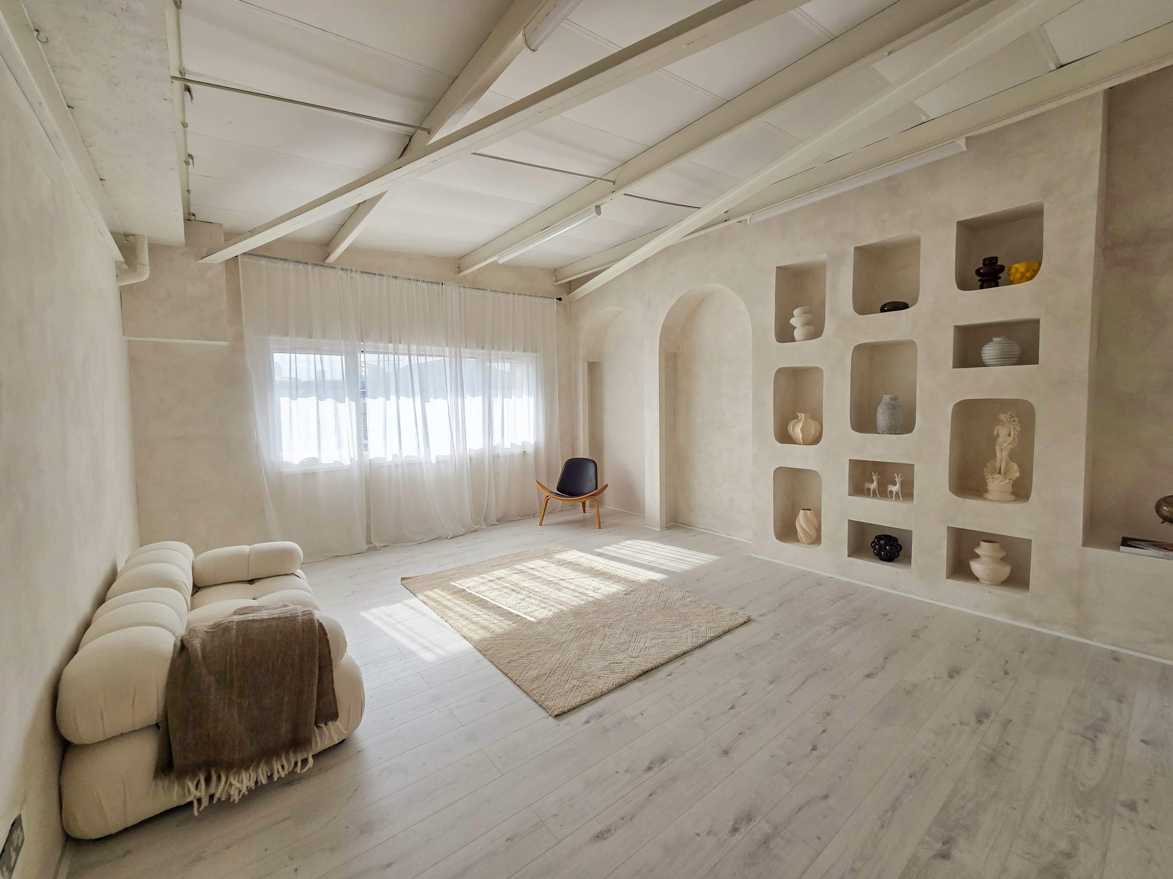 The image shows a spacious, minimalist room with light-colored walls, a textured rug on the wooden floor, a single chair, and a set of recessed shelves displaying various decorative items.