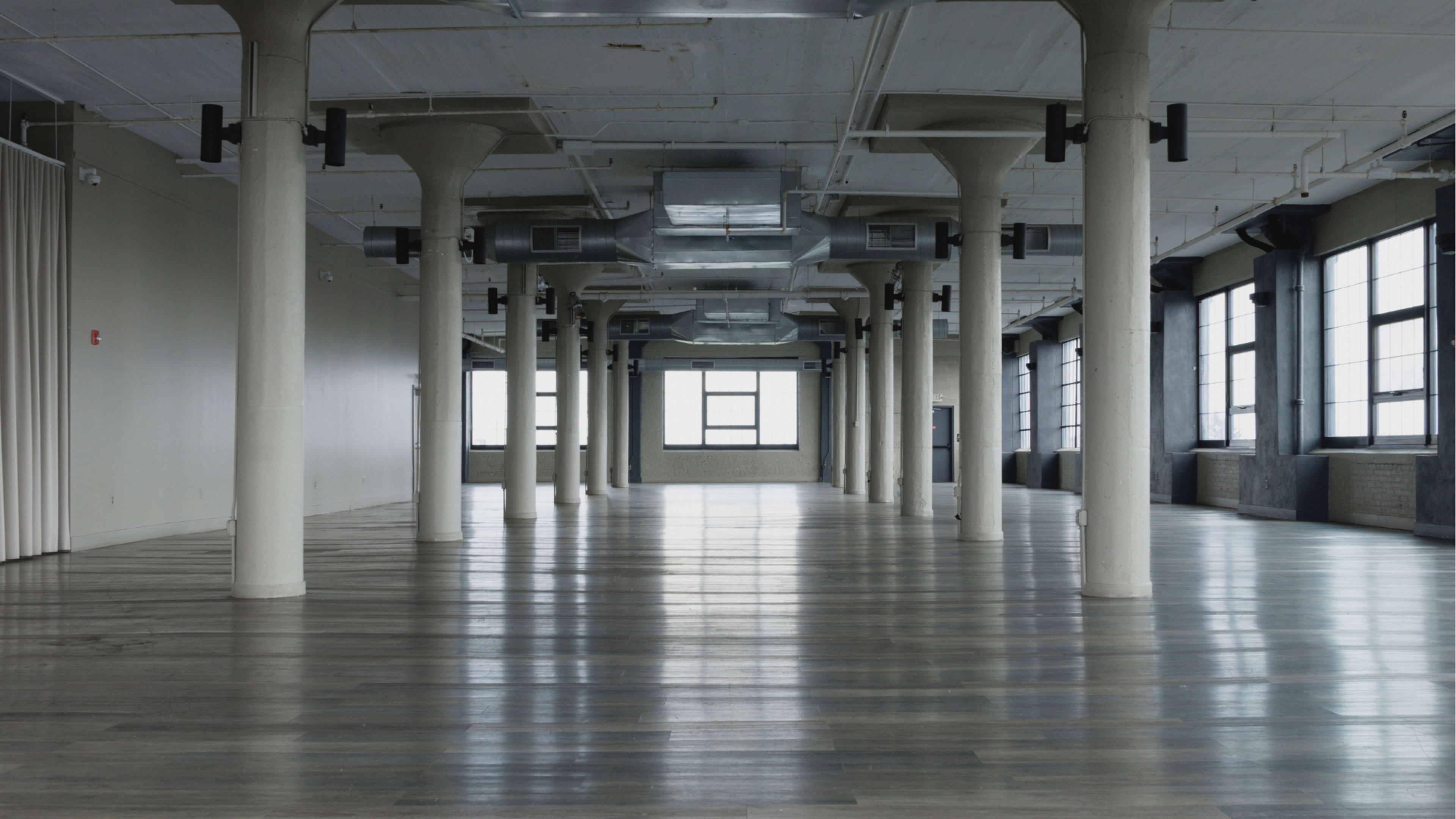 The image shows an empty, spacious room with large windows and supportive columns, featuring polished wooden floors and bare walls.