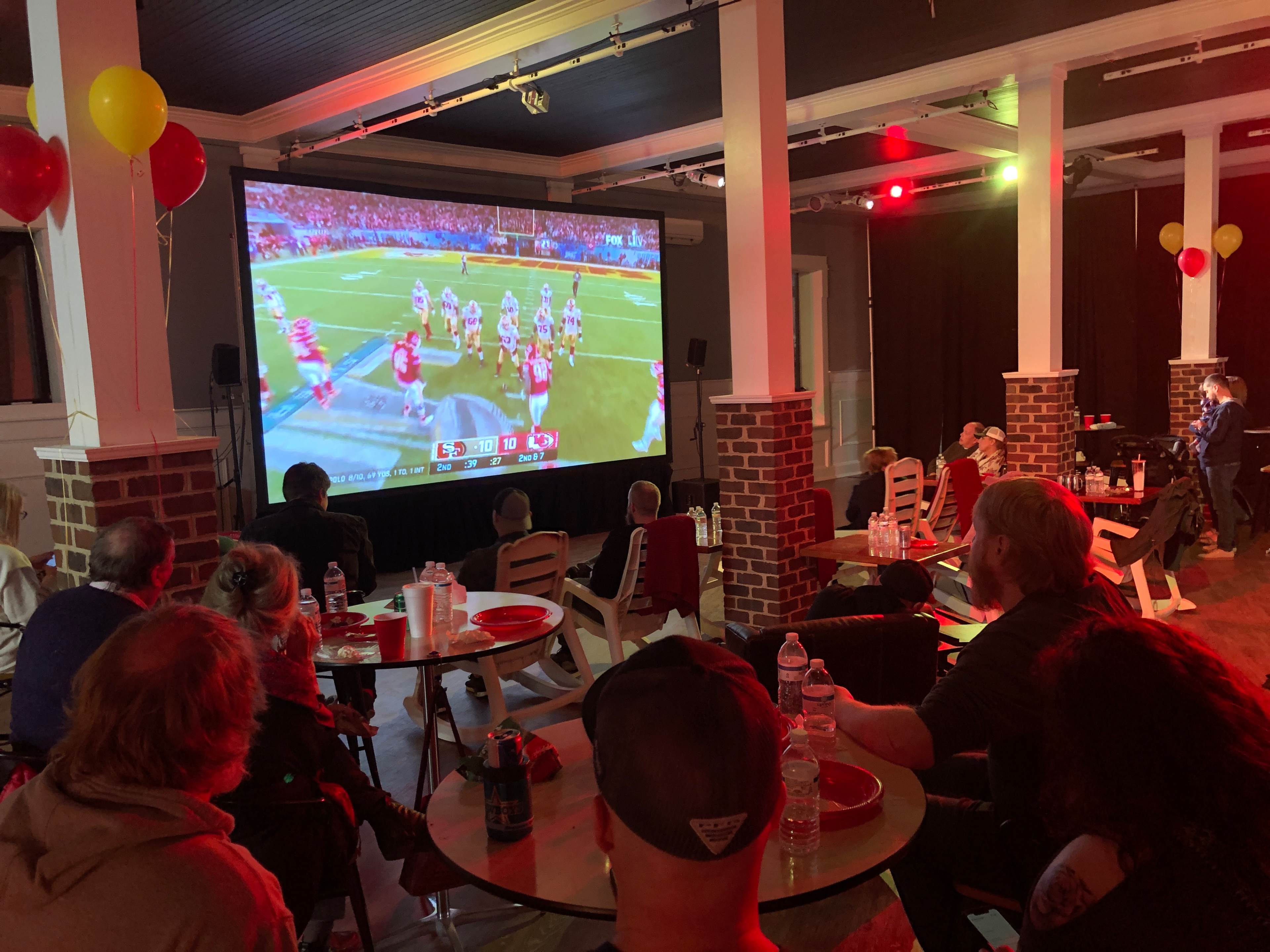 A group of people watches a football game on a large screen in a venue decorated with balloons and tables.