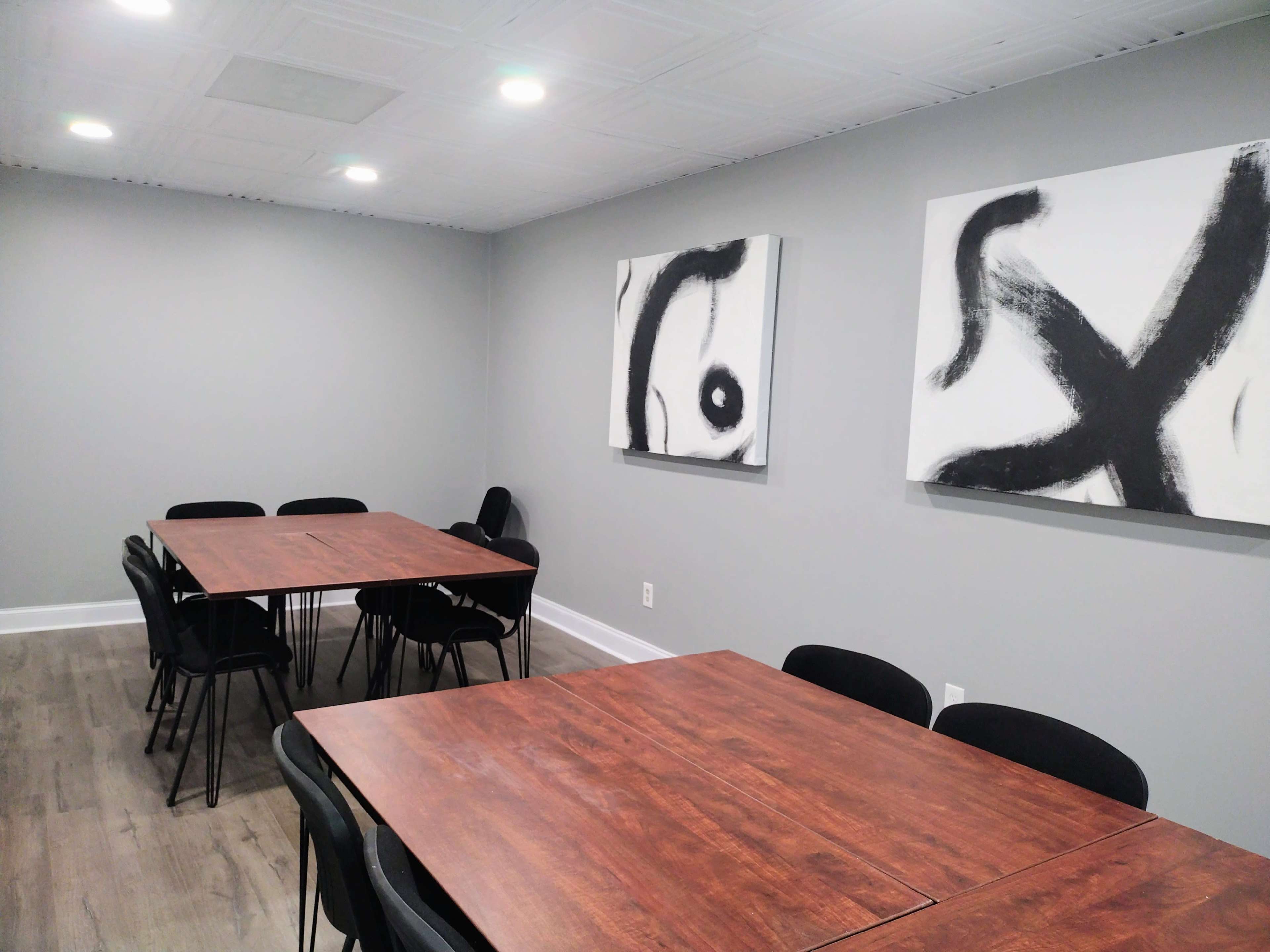 The image shows a meeting room with two large abstract paintings on the gray walls and four wooden tables surrounded by black chairs.