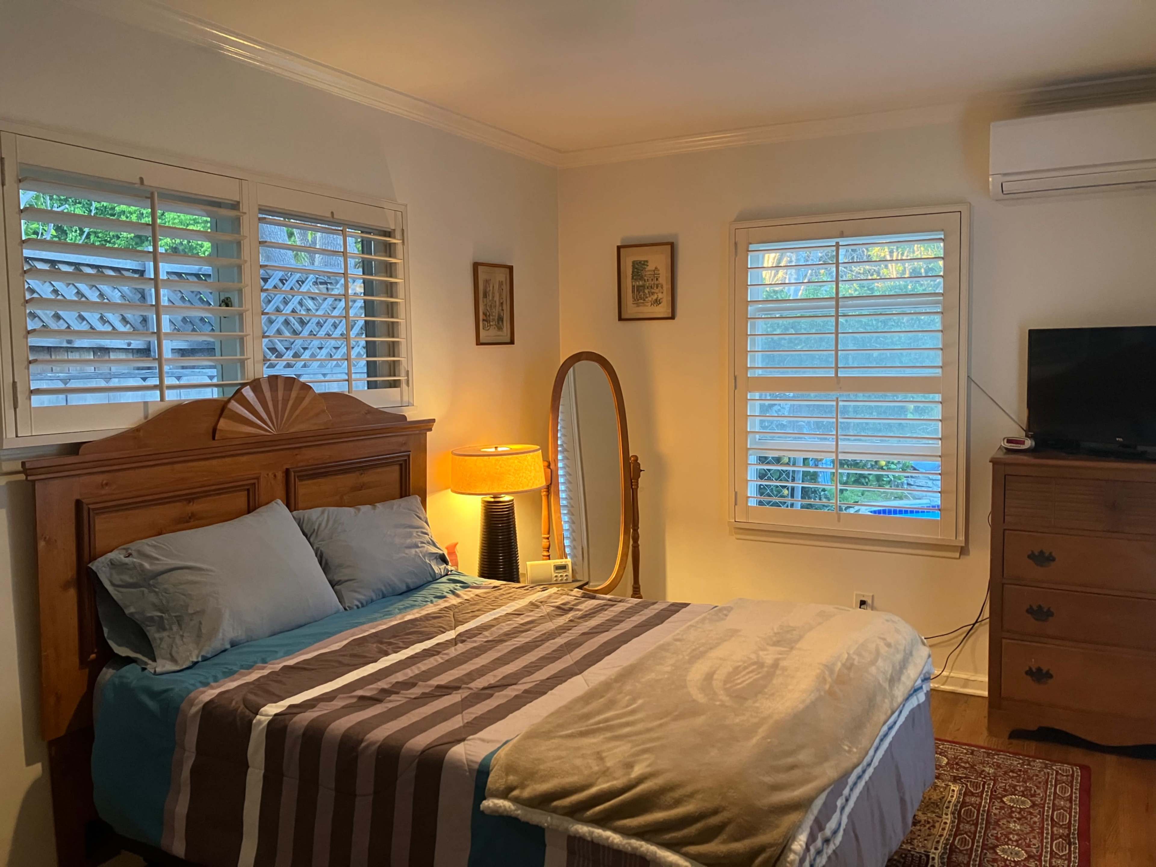 The image shows a cozy bedroom featuring a bed with blue and gray bedding, a wooden dresser, a tall mirror, a lamp, and two windows allowing natural light.
