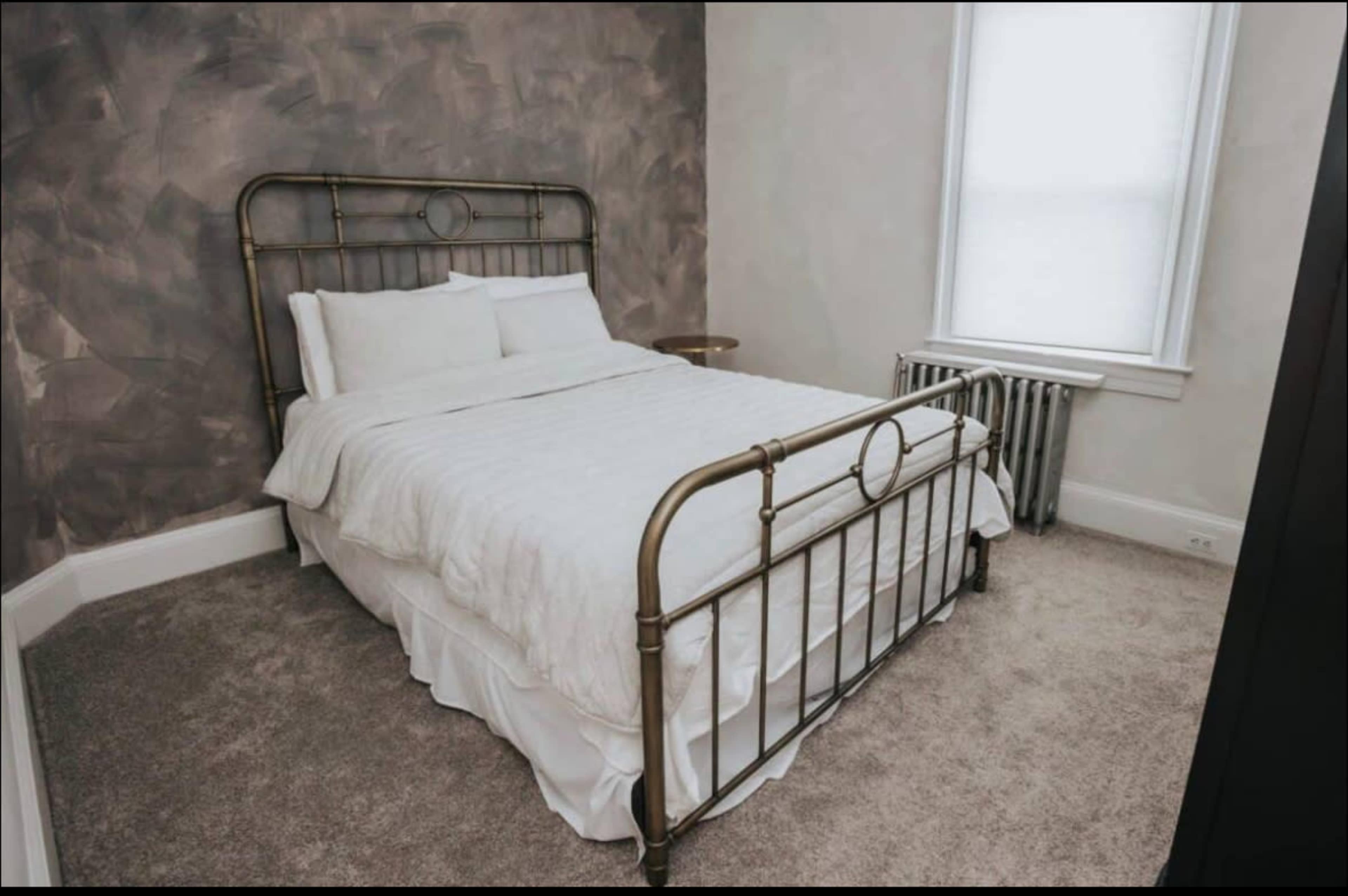 The image shows a neatly made bed with a brass frame positioned in a sparsely furnished bedroom with muted walls and a radiator.