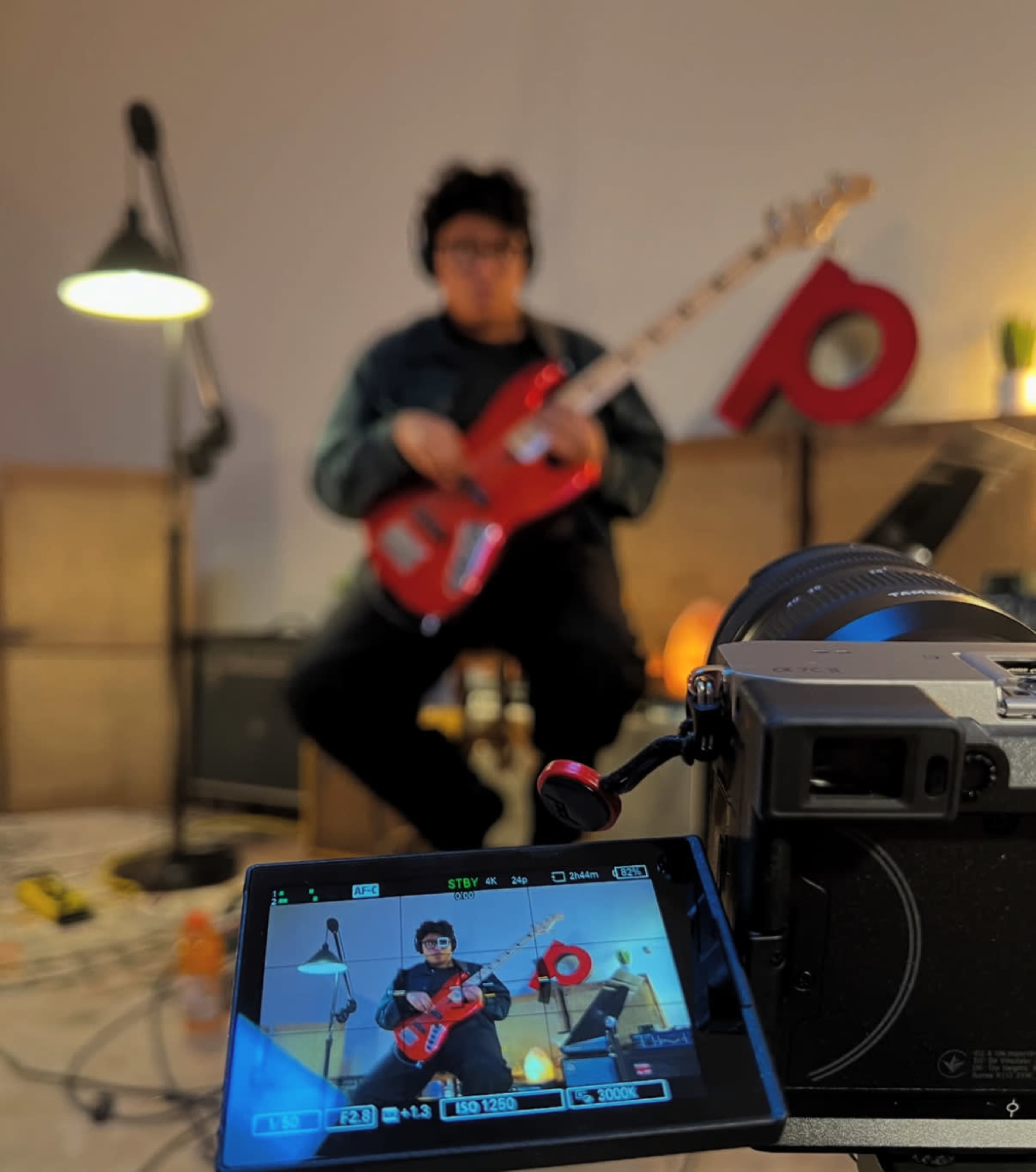 A person is sitting with a red guitar, while a camera displays their image on a screen in a dimly lit room with a lamp and decorations in the background.