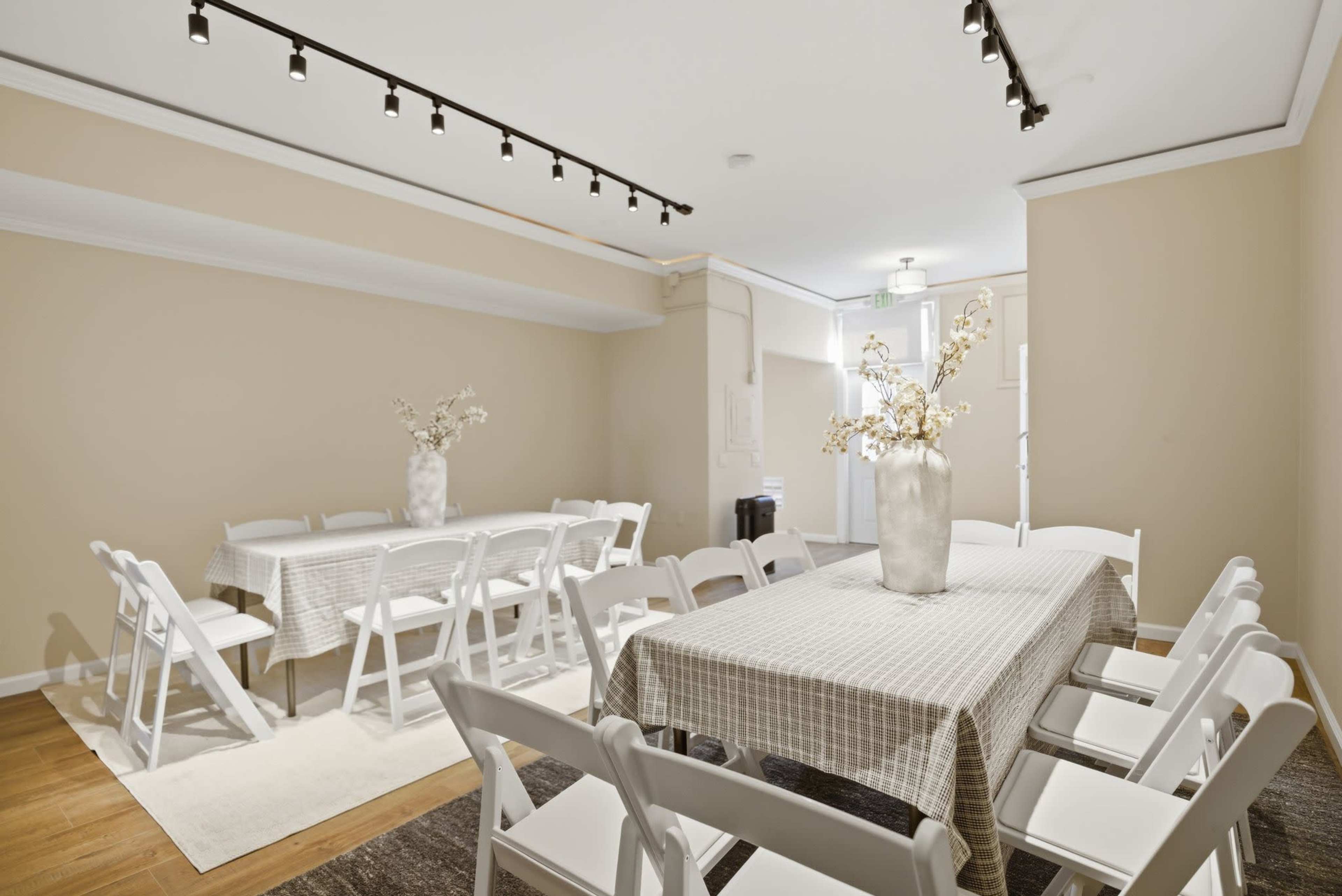 A spacious meeting room features several tables covered with checkered tablecloths and surrounded by white folding chairs, with a vase of flowers as the centerpiece.