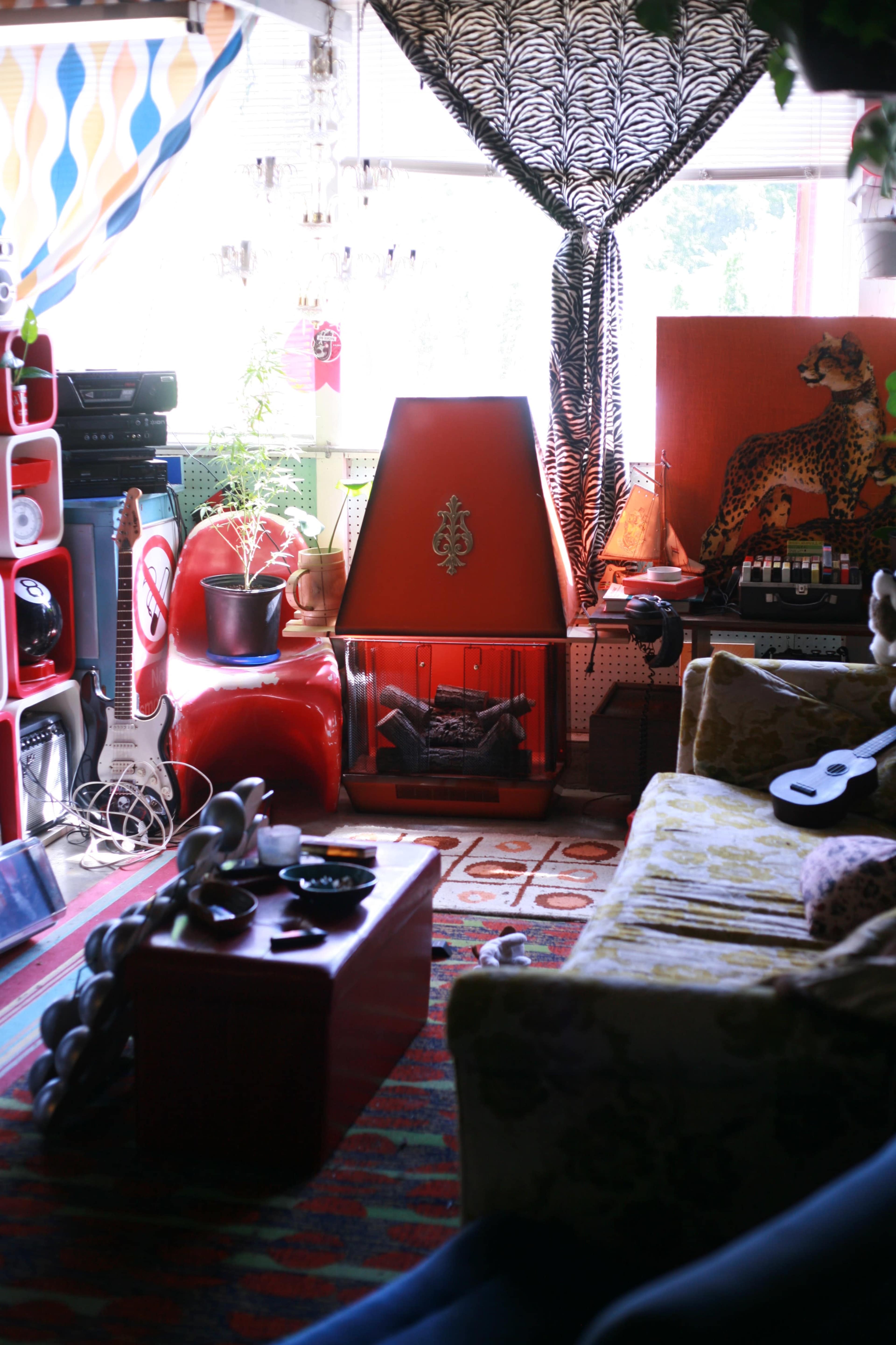 A colorful living room filled with eclectic furniture, a large window with patterned curtains, and a retro-style orange heater.