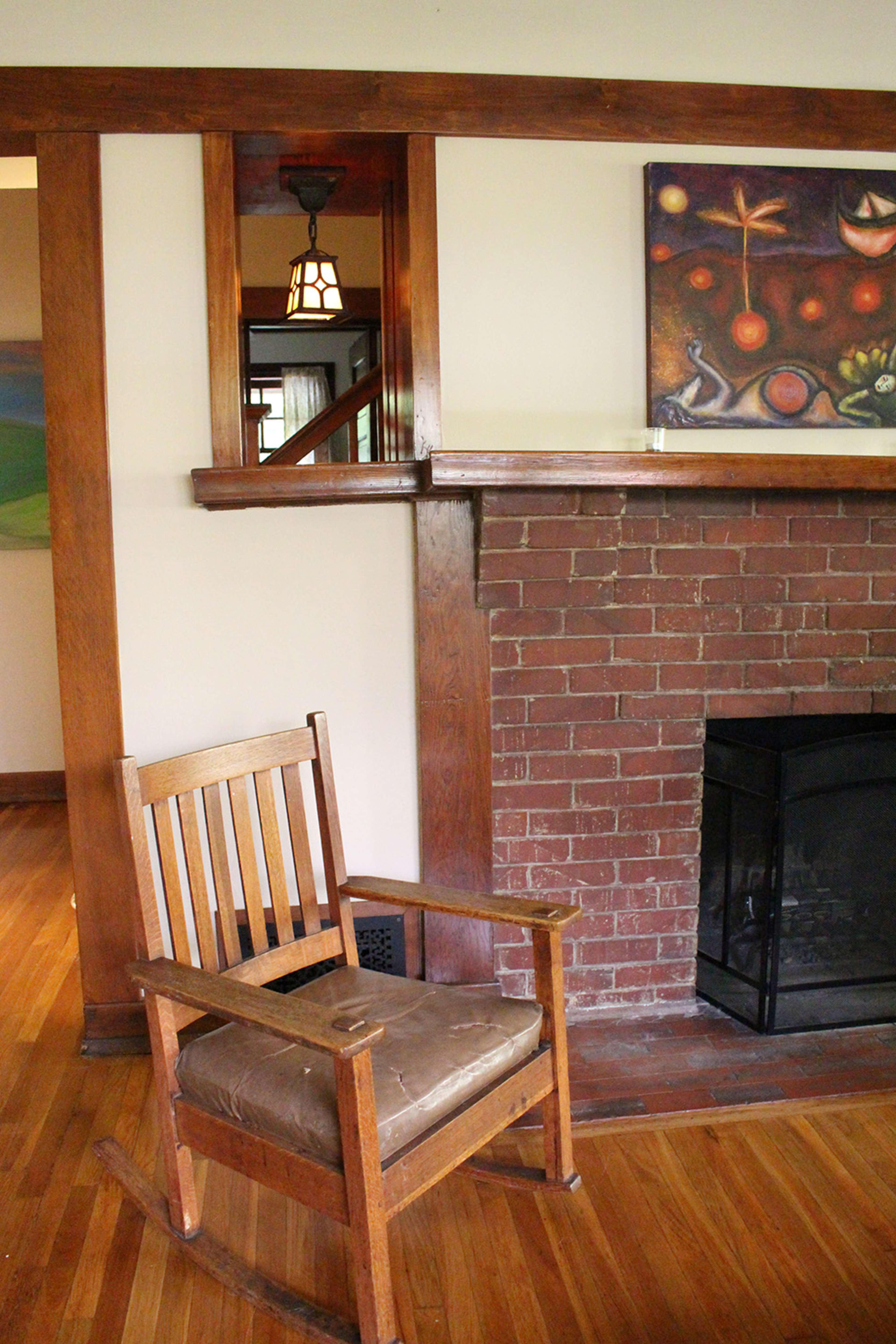 A wooden rocking chair sits in front of a brick fireplace in a room with wooden beams and artwork on the wall.