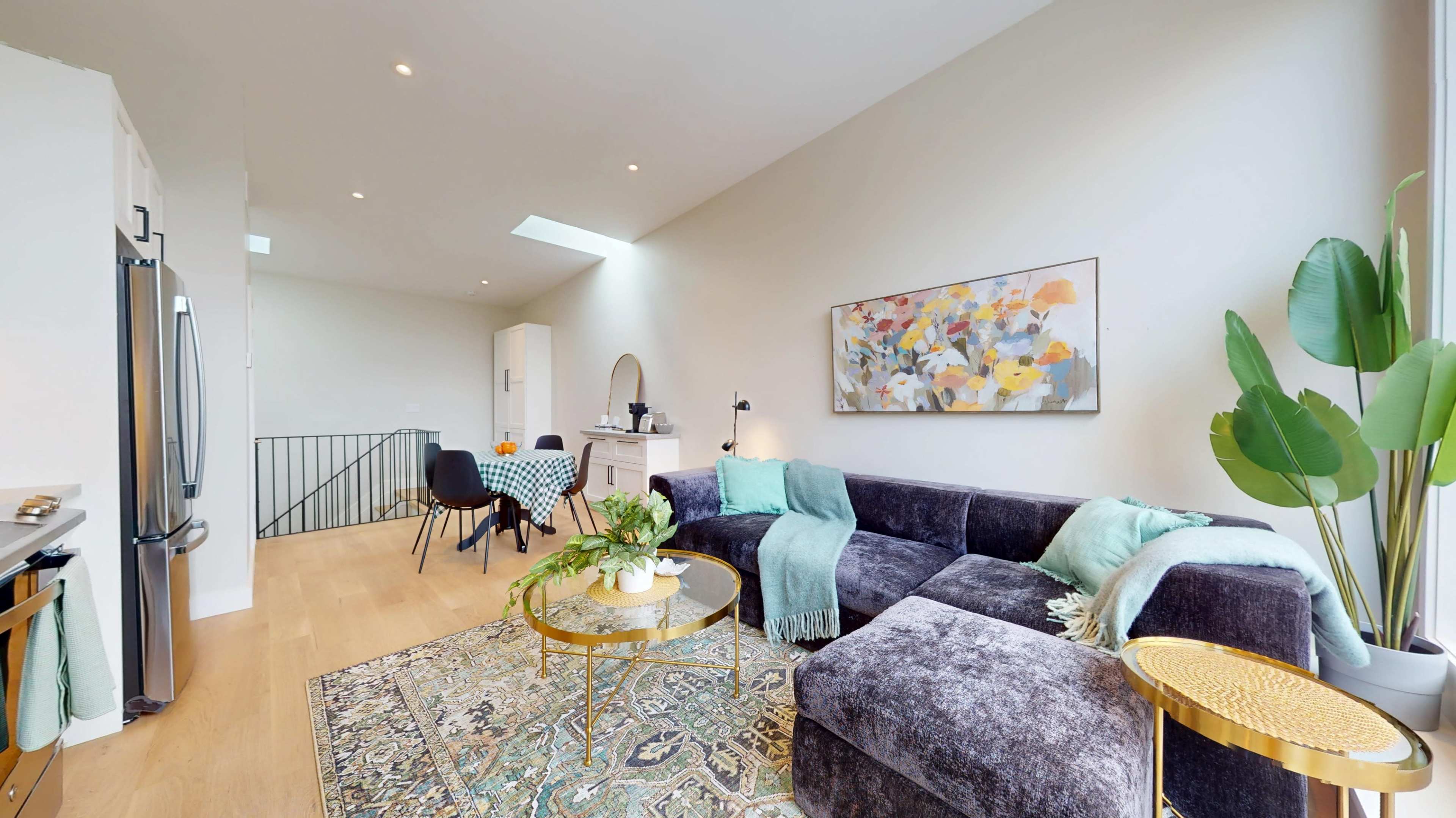 The image shows a modern living space featuring a dark gray sectional sofa, a round gold coffee table, a decorative area rug, and a dining table set for four.